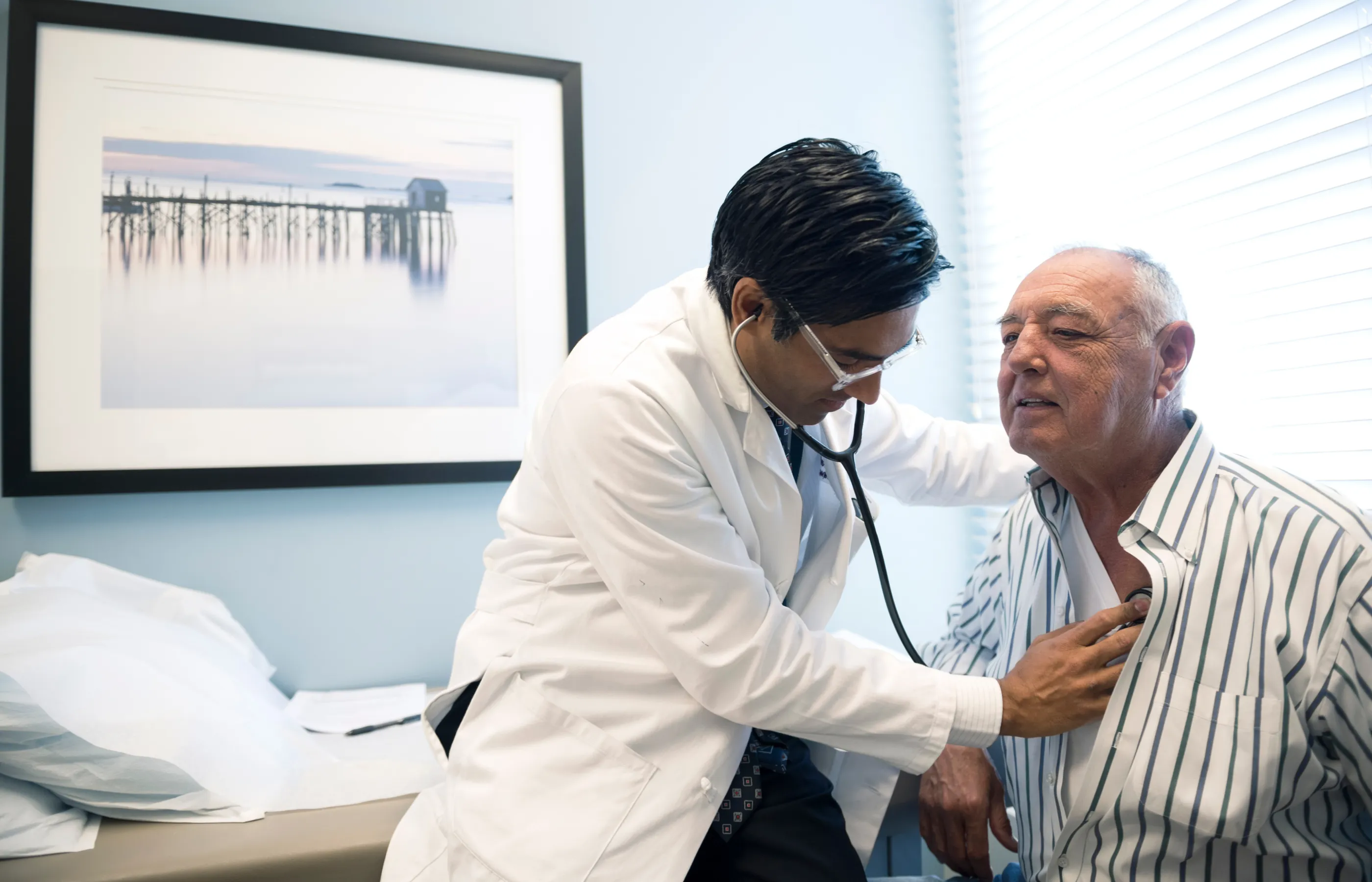 Dr. Theuruvath is leaning in to listen to a senior male patient heartbeat with a stethoscope. 