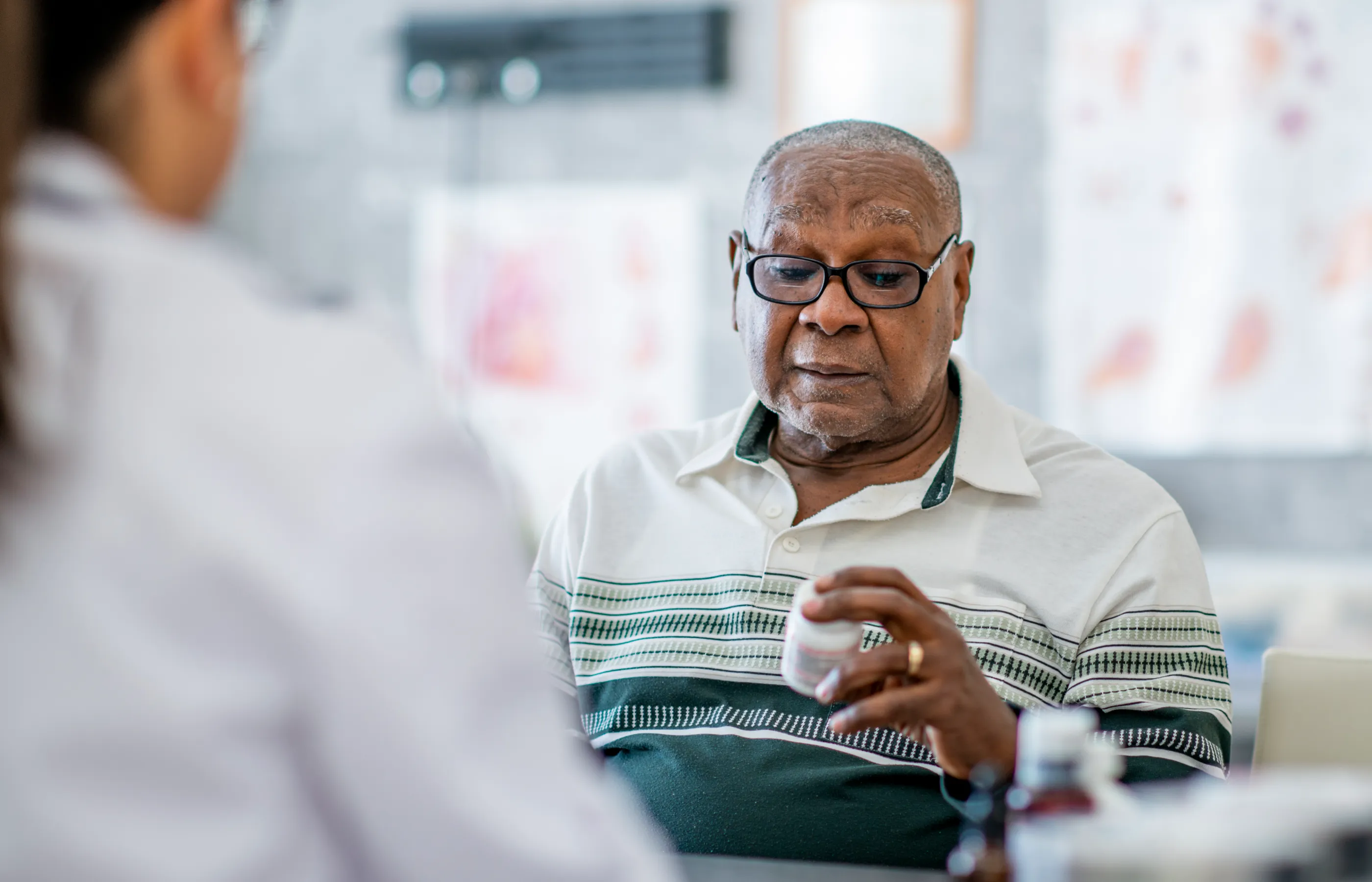 An elderly man looks at a pill bottle in his hand while sitting across the table from a doctor in the doctor's office.