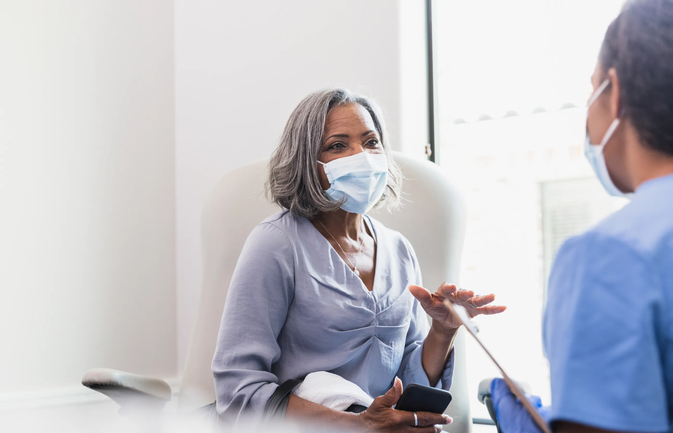 A senior woman is sitting with a mask and smart phone in her hand as she talks with her doctor in an exam room. 