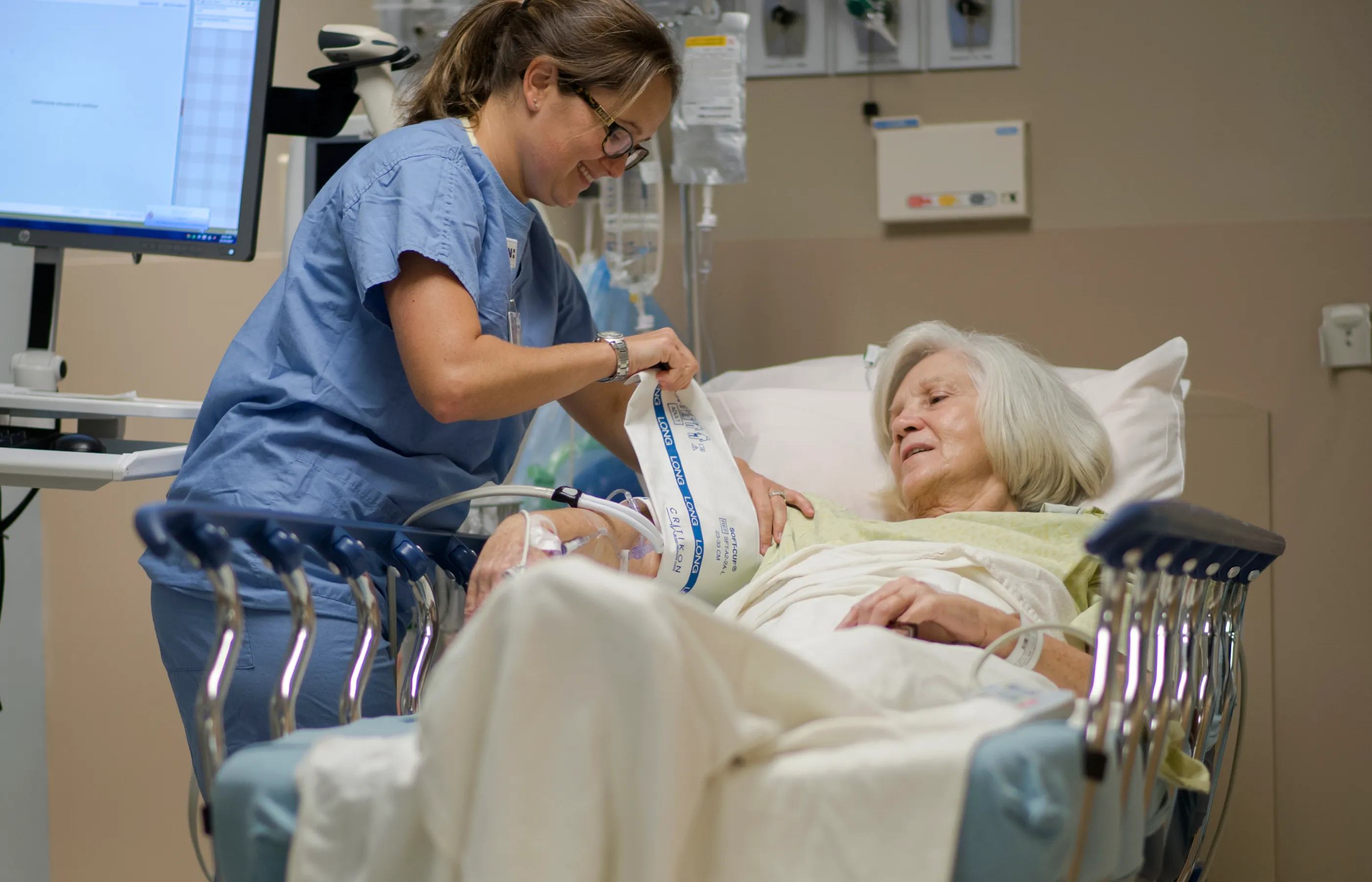 Nurse and patient are in the post-op area of the hospital. The patient is lying in bed, while the nurse wraps a blood pressure cuff around her arm.  