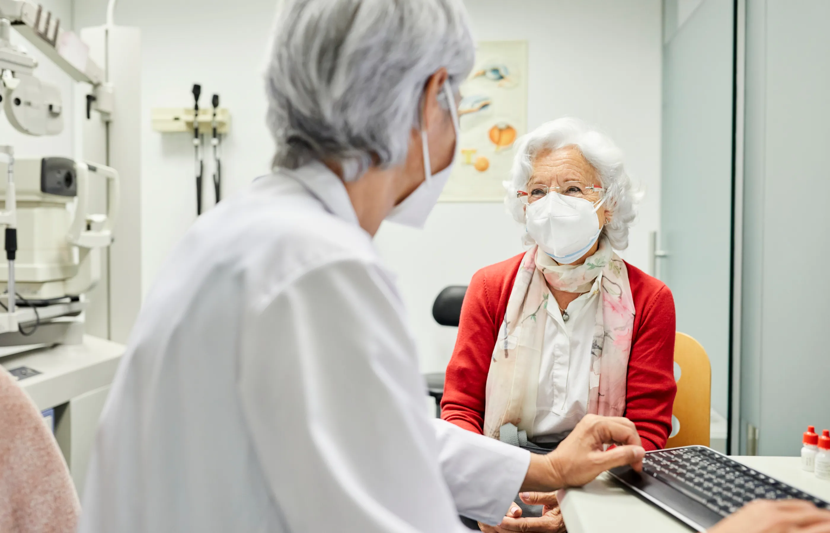 A senior woman and her doctor are sitting in an exam room talking to each other. Off to the side, you see a Phoropter, which measure refraction for vision correction. 