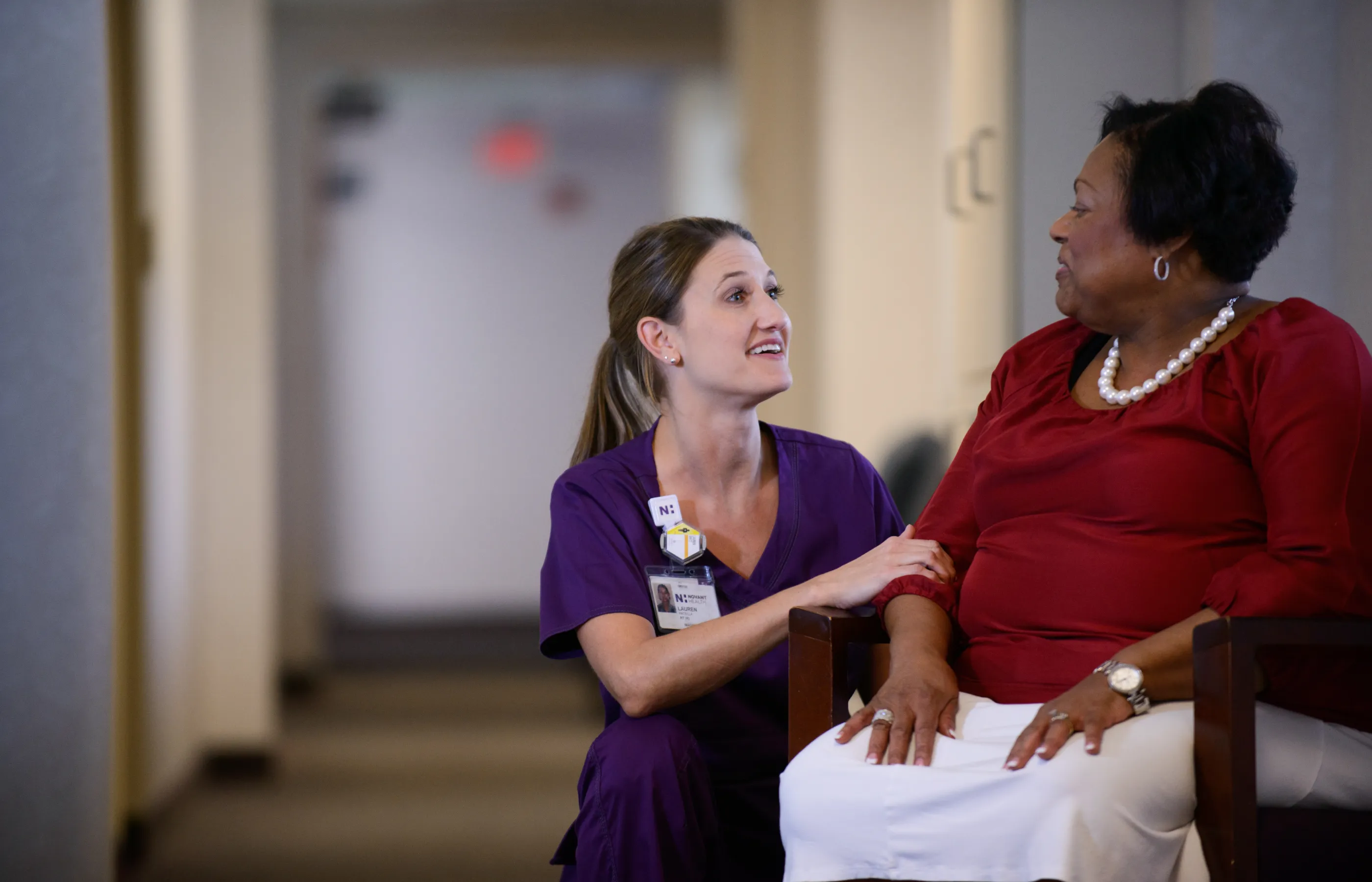 A Novant Health team member is kneeling next to a patient as she talks with her. The patient is looking down and smiling at the team member. 