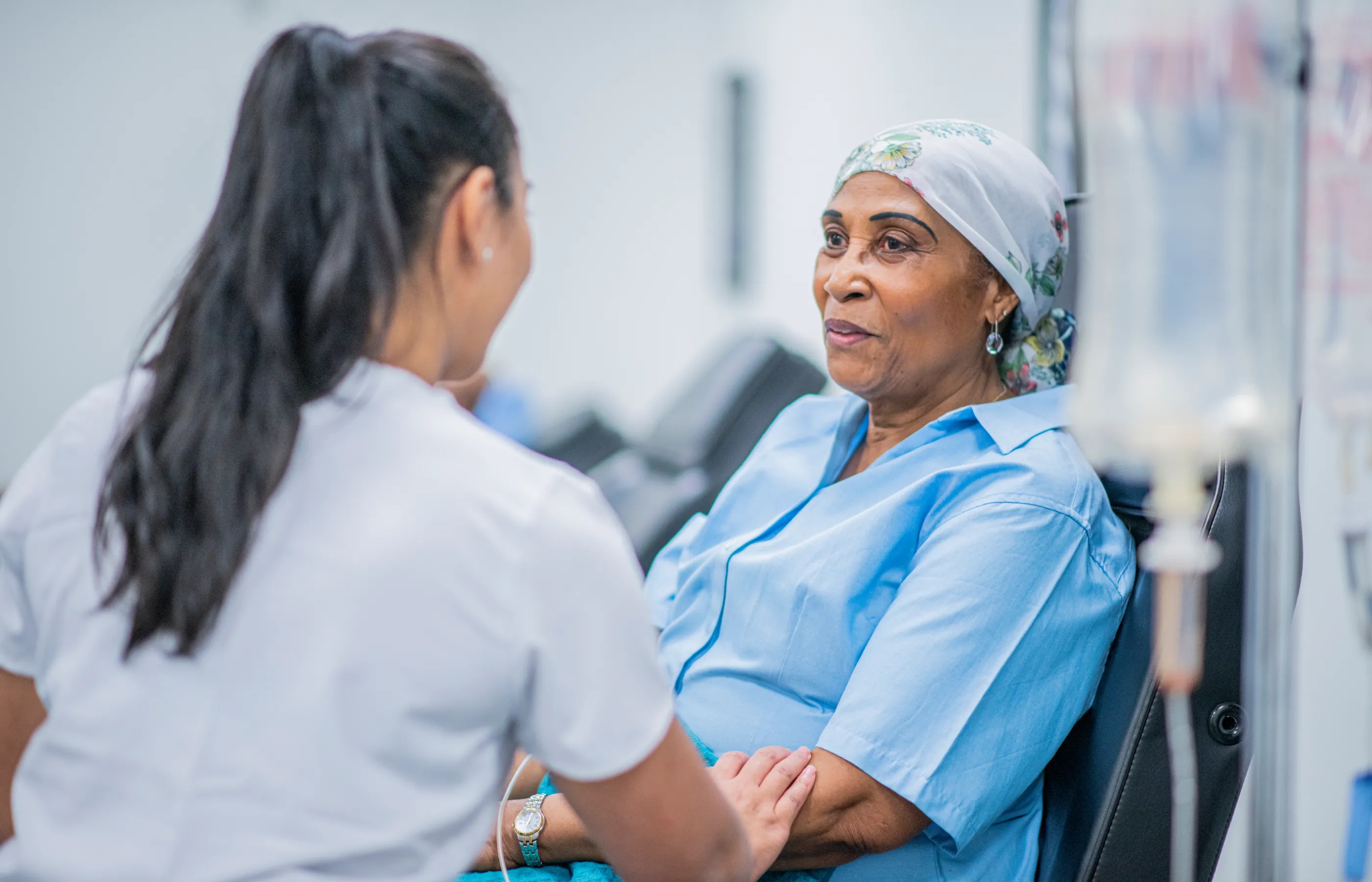 A woman is undergoing cancer treatment as a nurse stands in front of her. In support, the nurse rests her hand on the patients arm. 