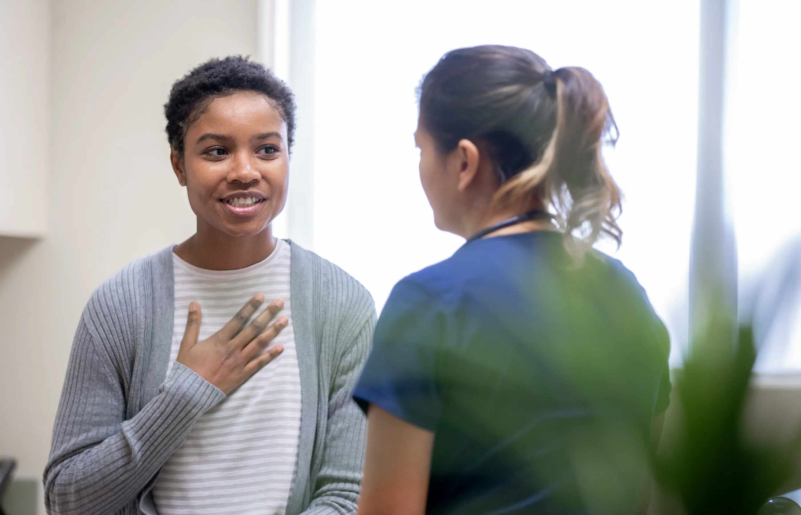 A young woman is sitting in an exam room talking with a nurse. As she talks, she has placed her hand over her chest. 