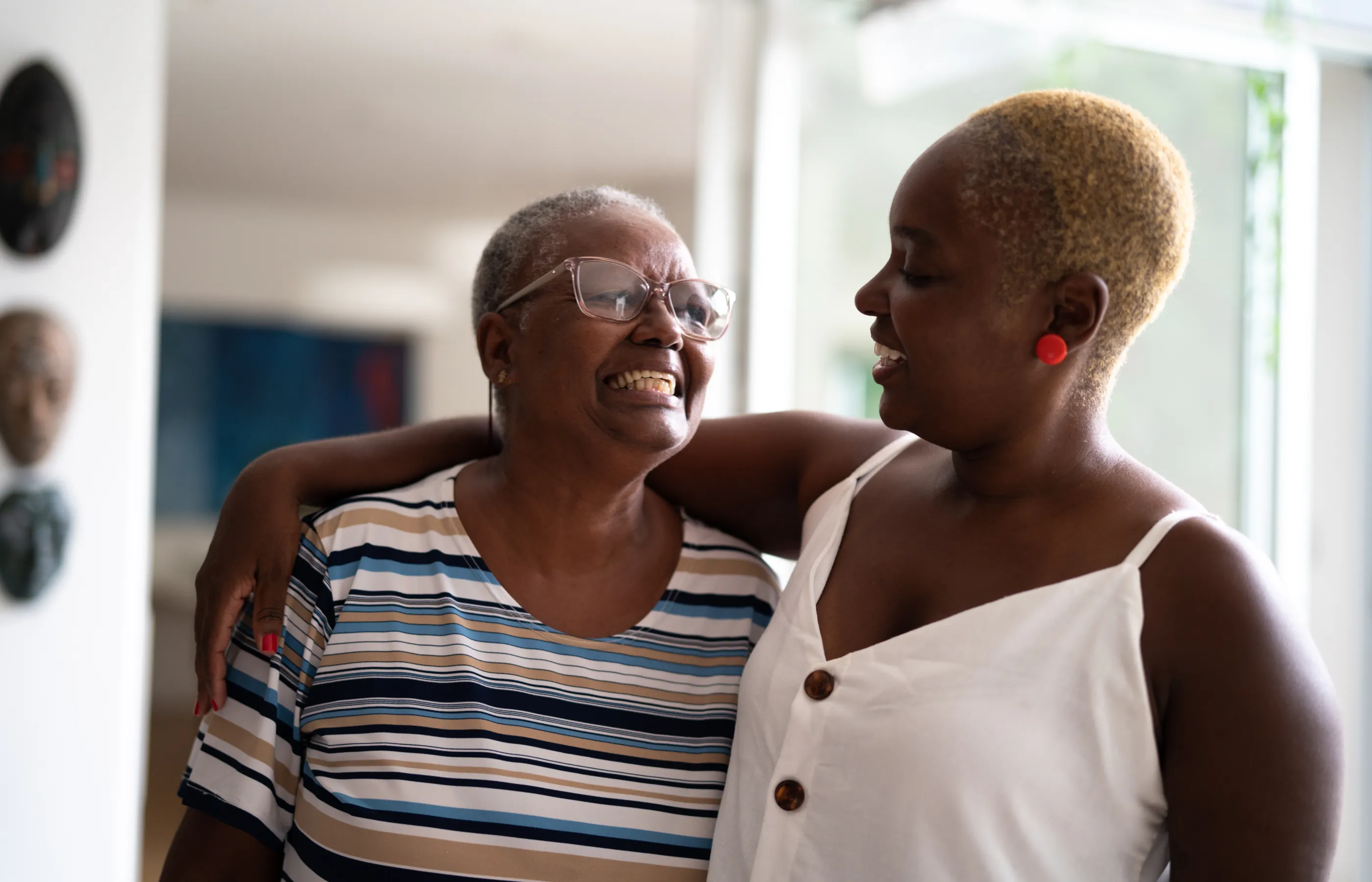 An adult woman is hugging and watching her mother smile in the hallway of her home. 