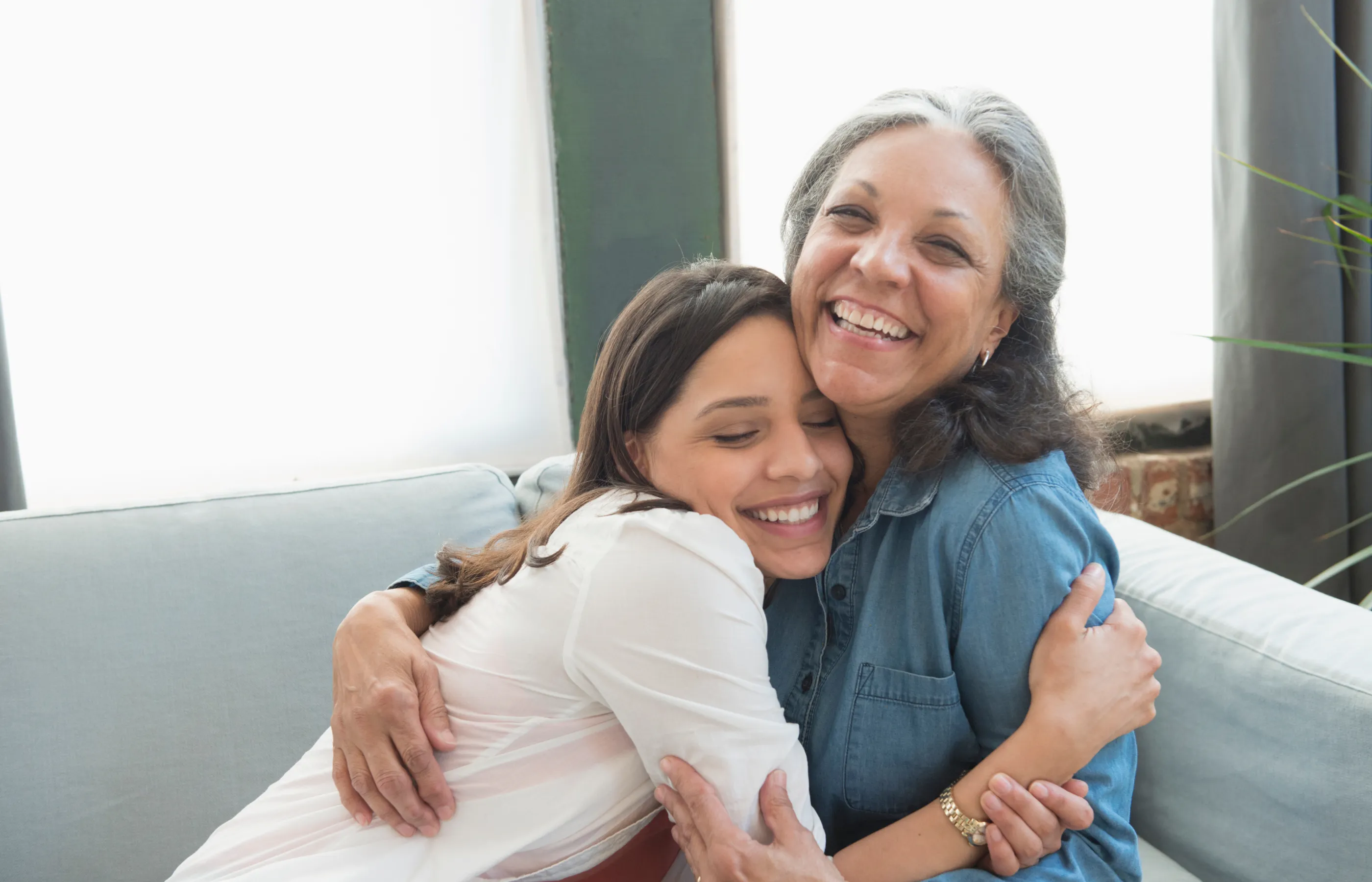 A mother is sitting with and hugging her adult child. They both smile as they hug each other. 