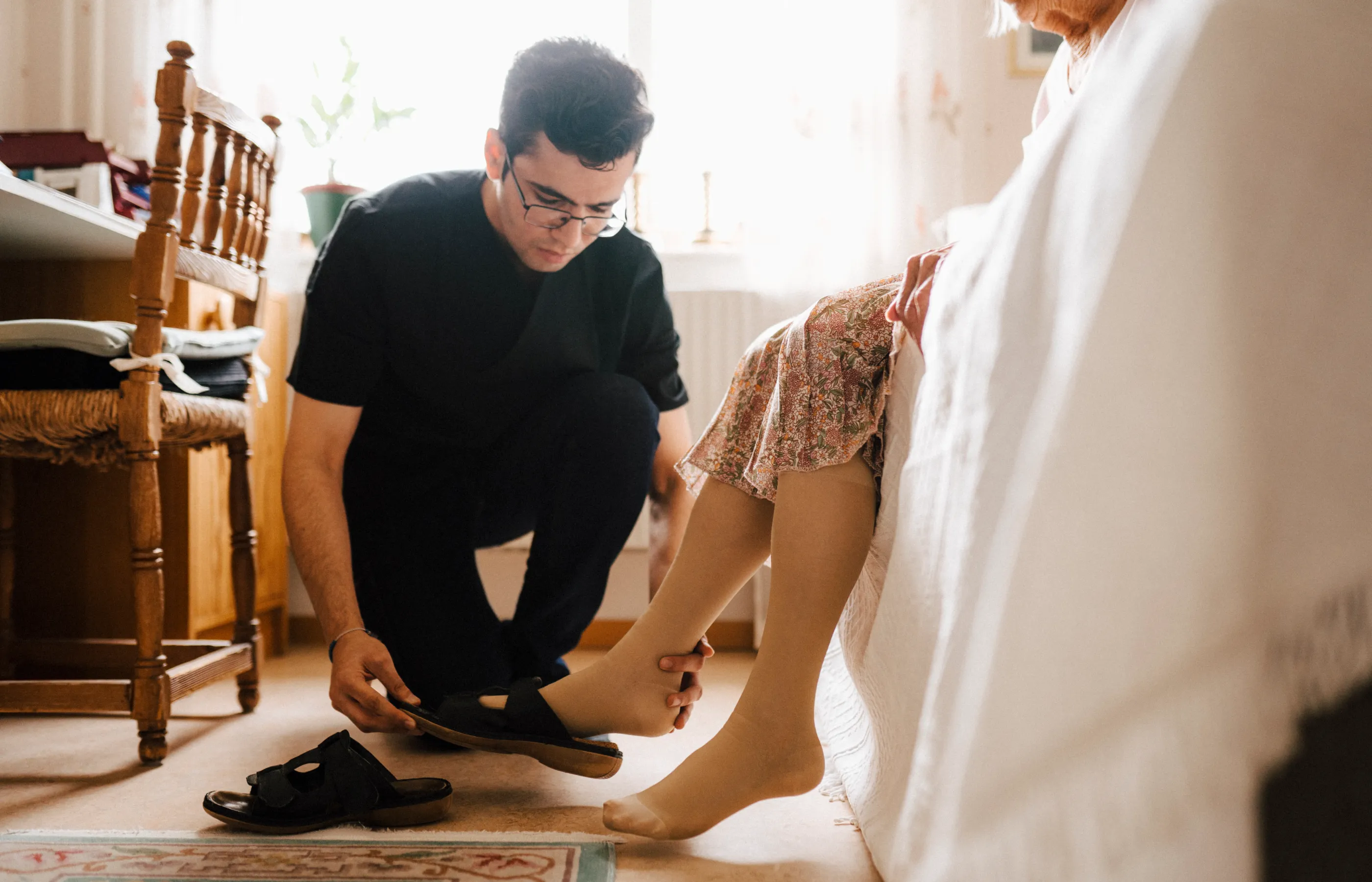 A caregiver is kneeling down to assist a senior woman by putting her shoes on. 