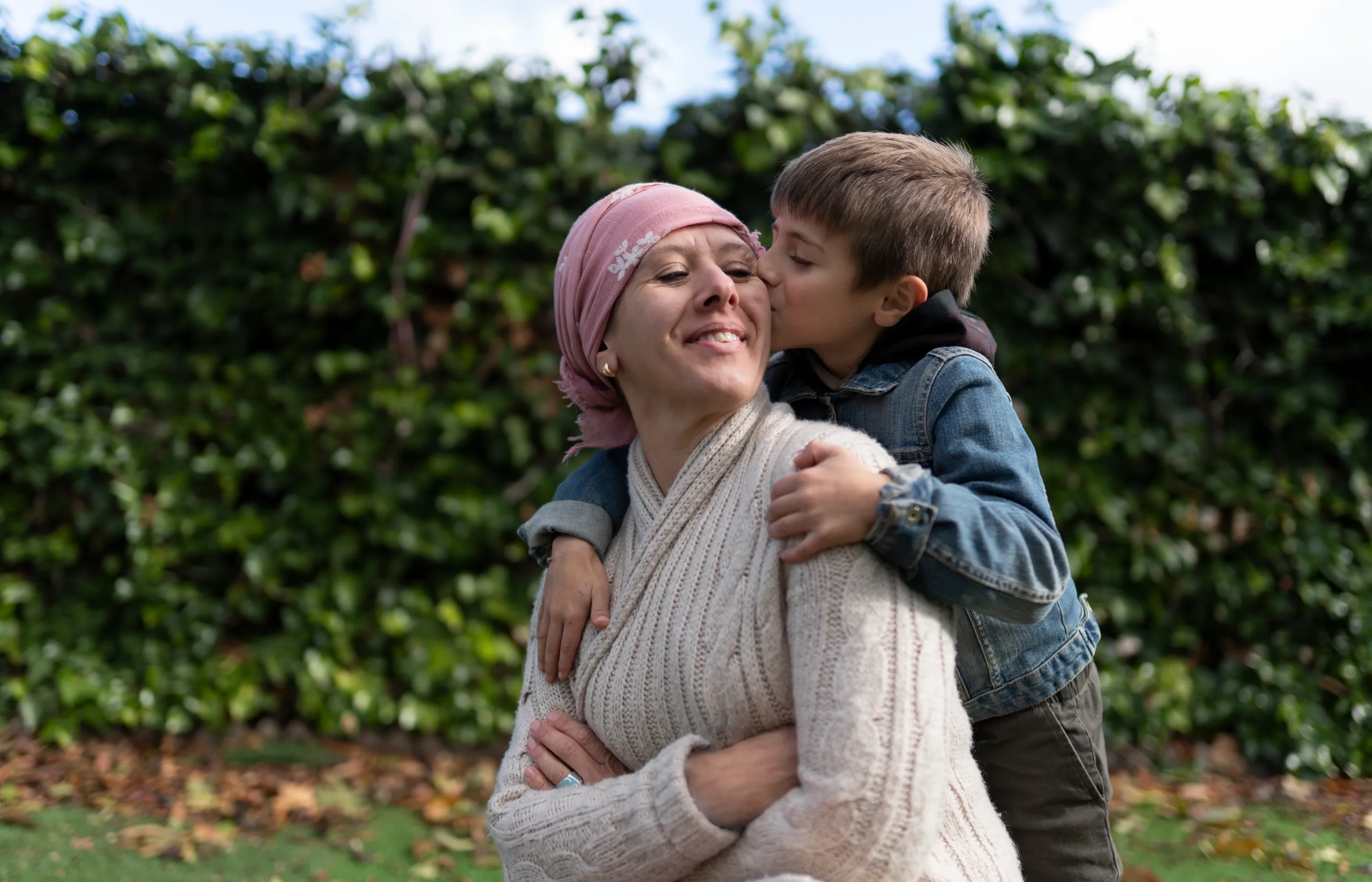 A cancer patient, is out in the back yard with her young child. the child is standing behind her, hugging, and kissing her cheek. 