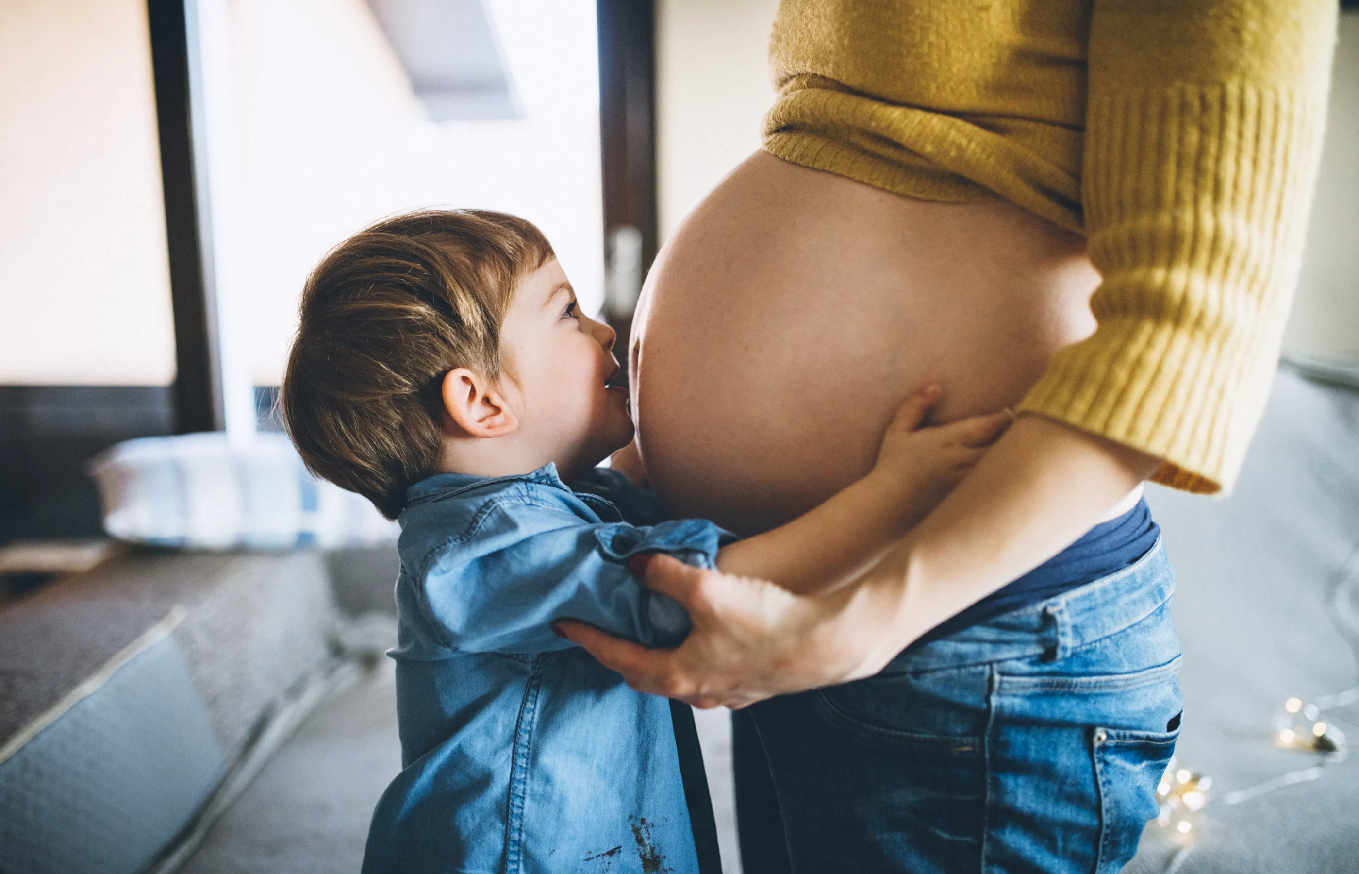 A pregnant woman is standing with her young child. The child is hugging and kissing his mother's baby bump. 