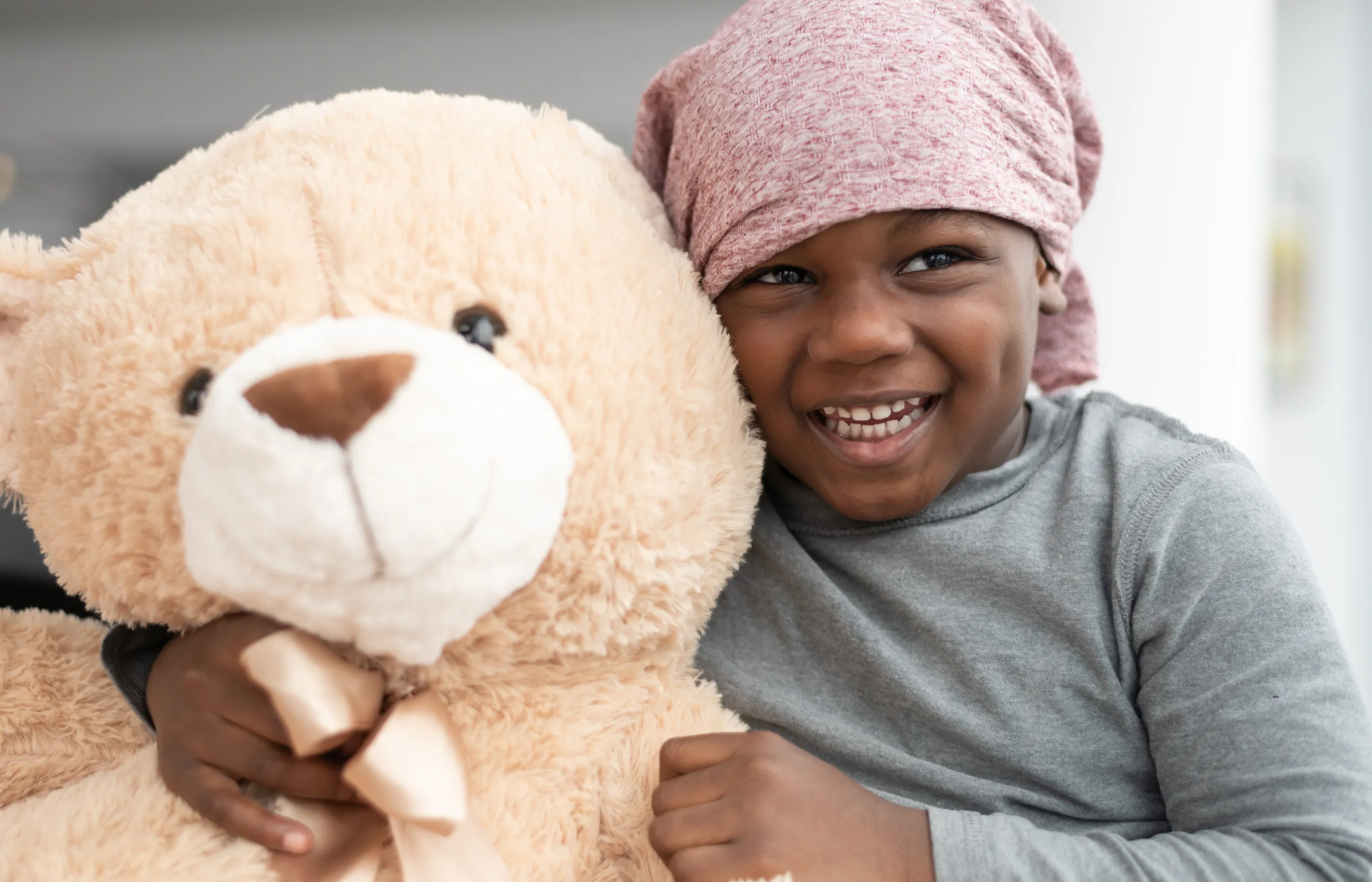 Young child wearing is wearing a head wrap and hugging a teddy bear. 