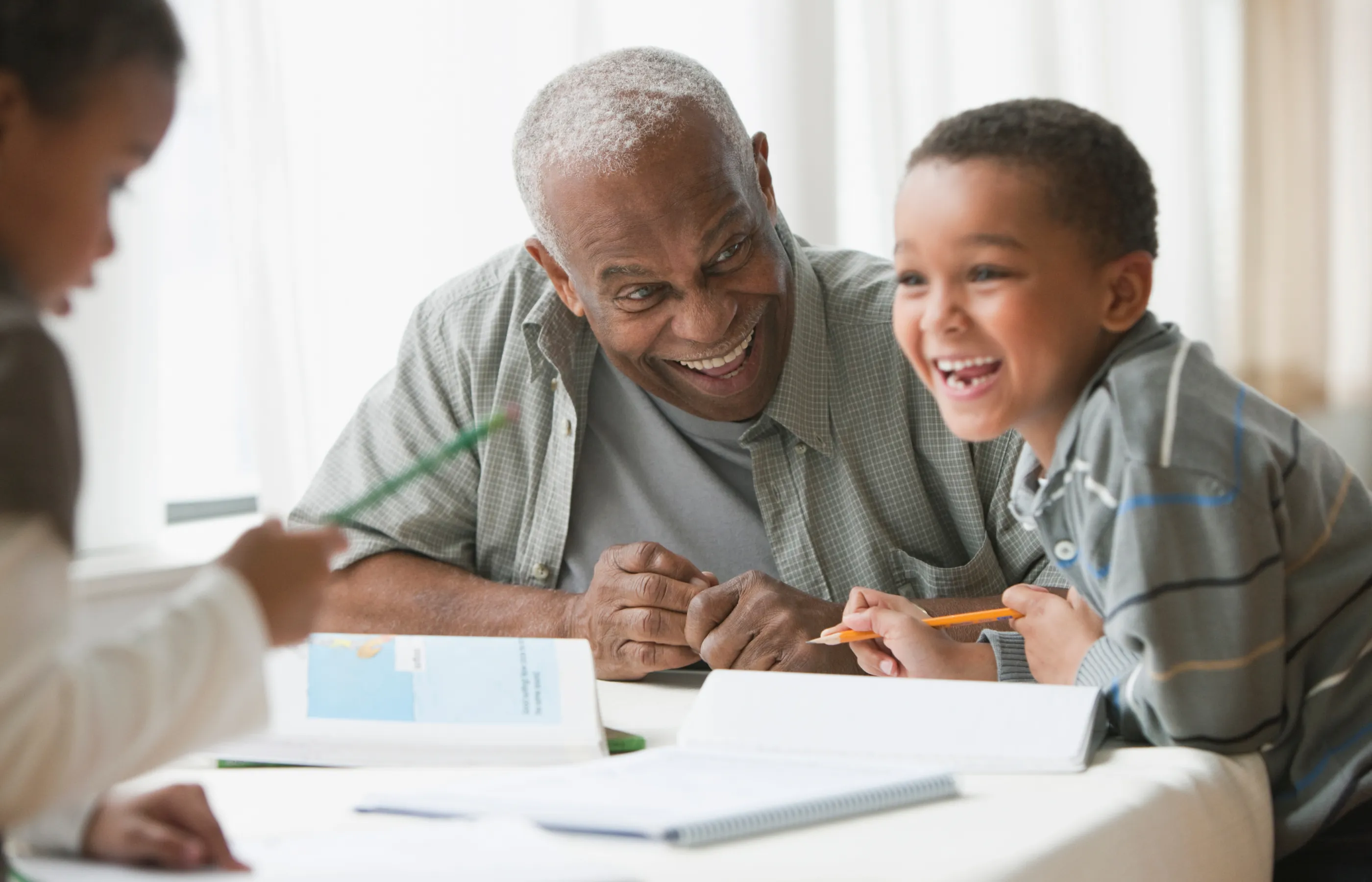 A grandfather with his two grandchildren. They are sitting at a table and smiling as they work on their homework. 