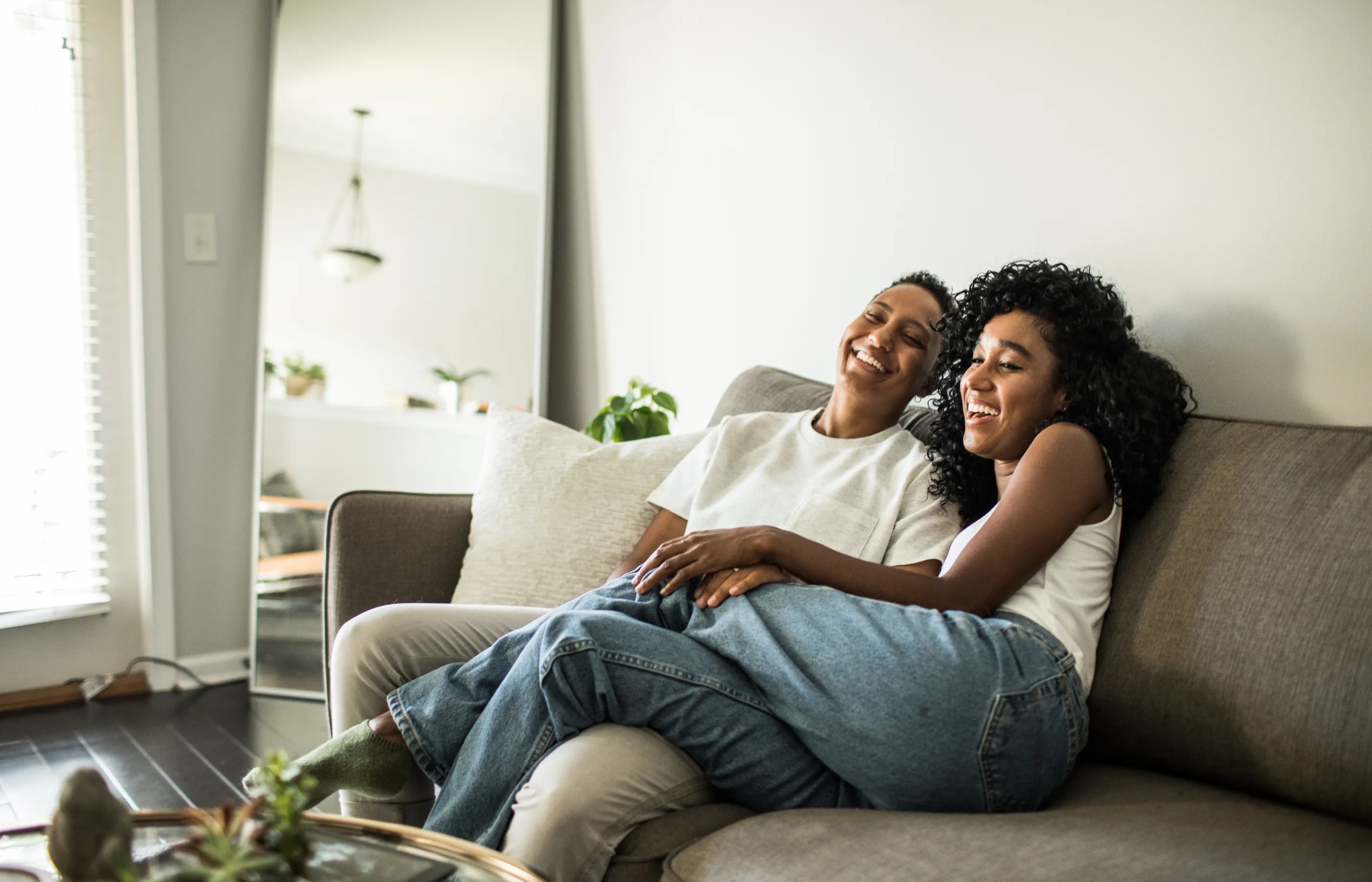 A lesbian couple is sitting on the couch in their home laughing. A lesbian couple is sitting on the couch in their home laughing.