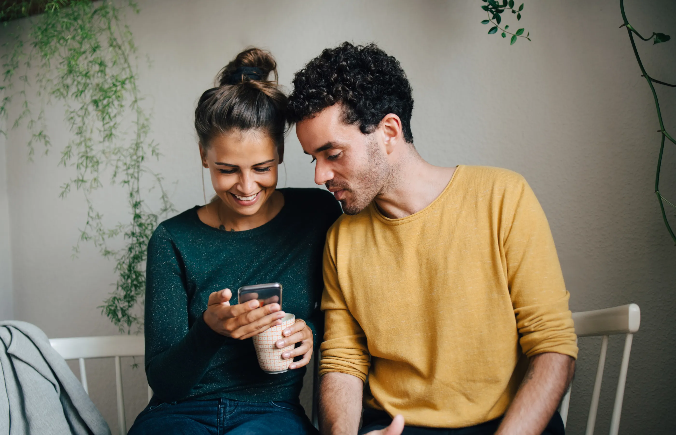 A couple is sitting on a bench together. The woman is holding a mug and smartphone. The man has leaned in close to look at the smartphone along with her.