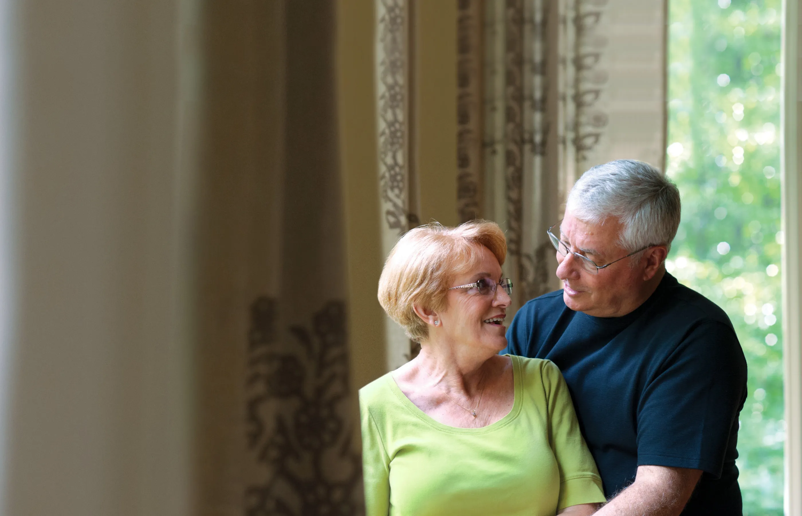 A senior man is standing behind his partner as he holds her hand. 