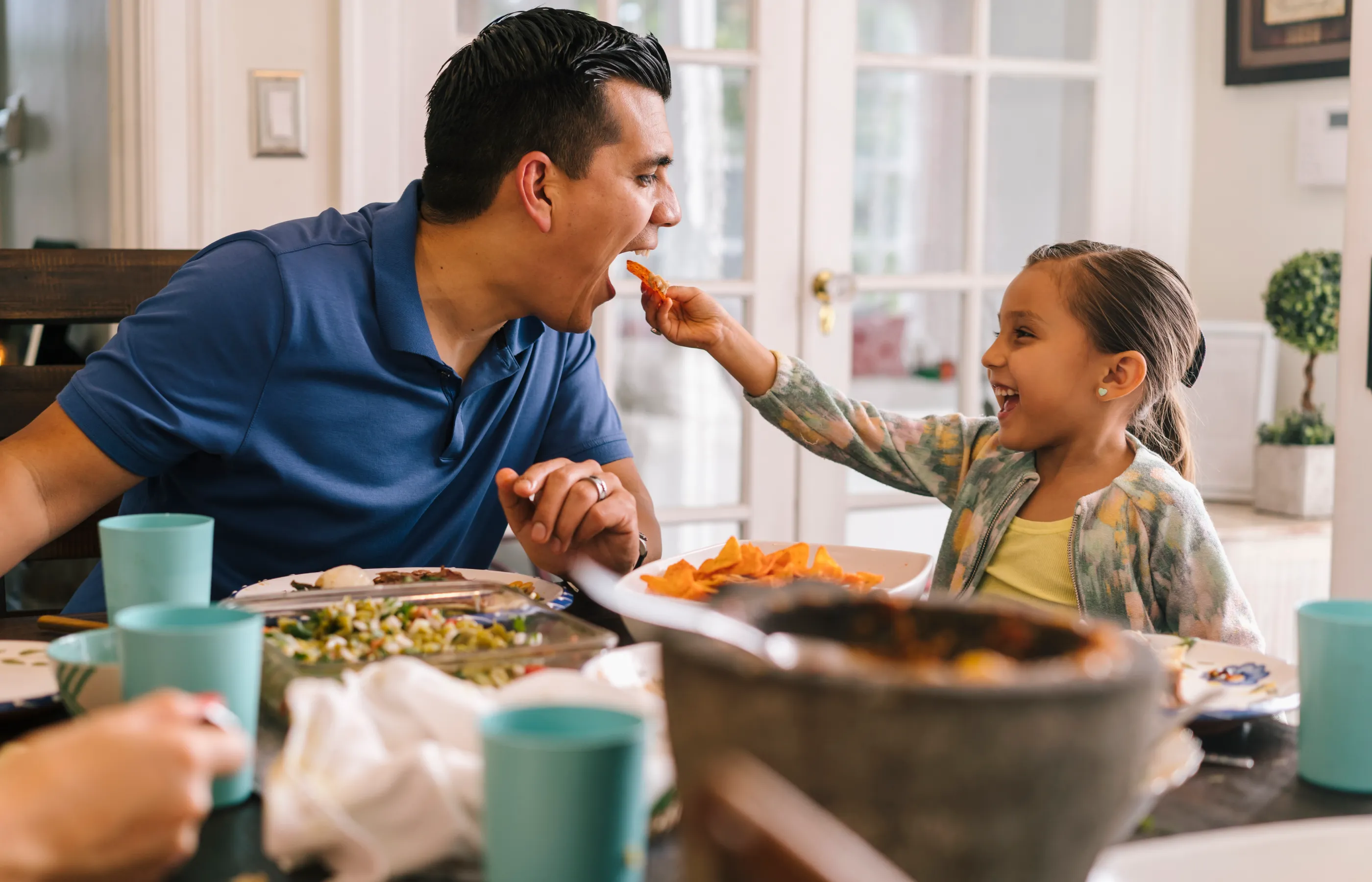 A young girl is laughing and feeding her dad food at the dinner table. 