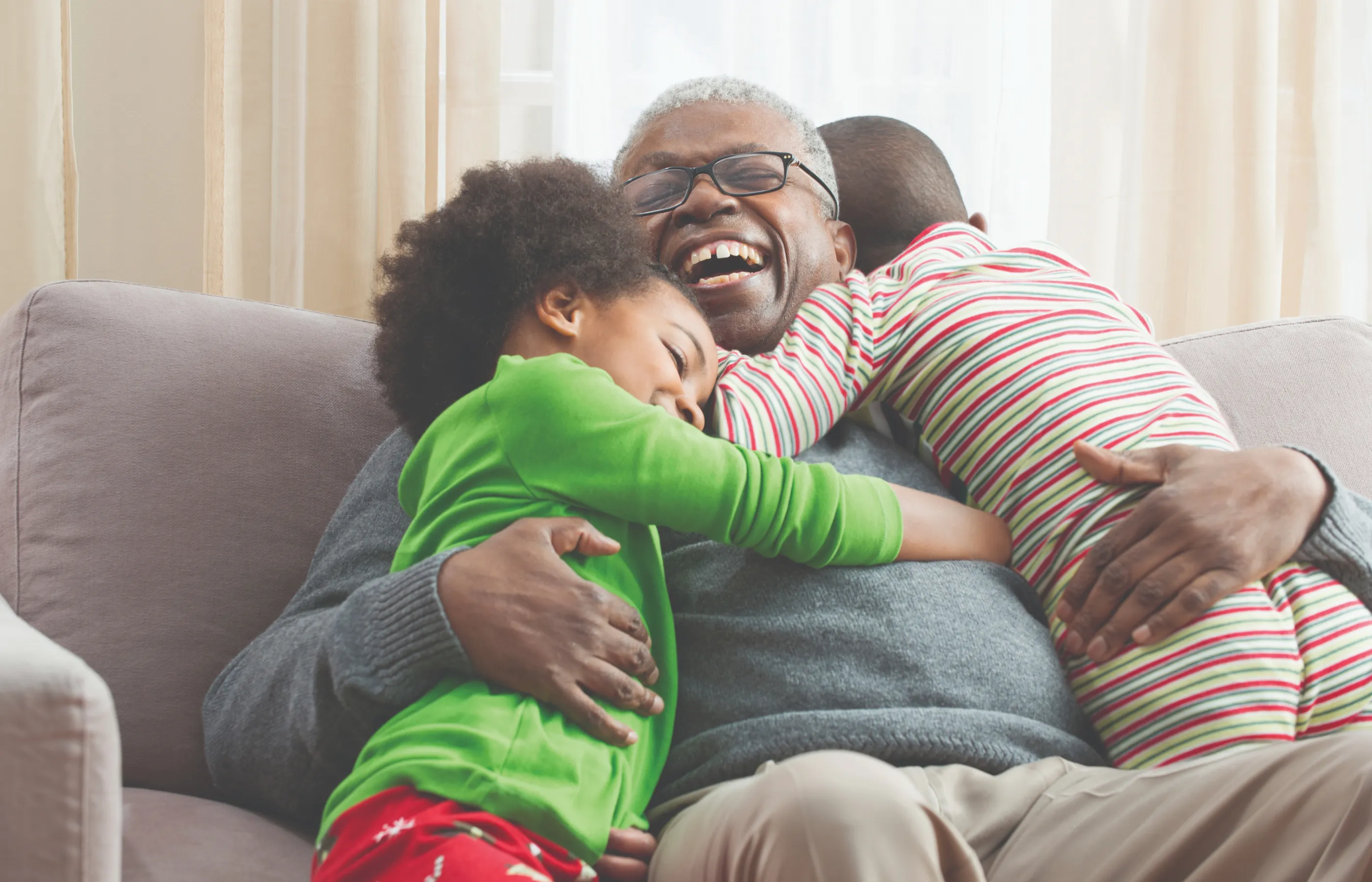 A grandfather is sitting on the couch embracing his two young grandchildren as they jump into his arms.