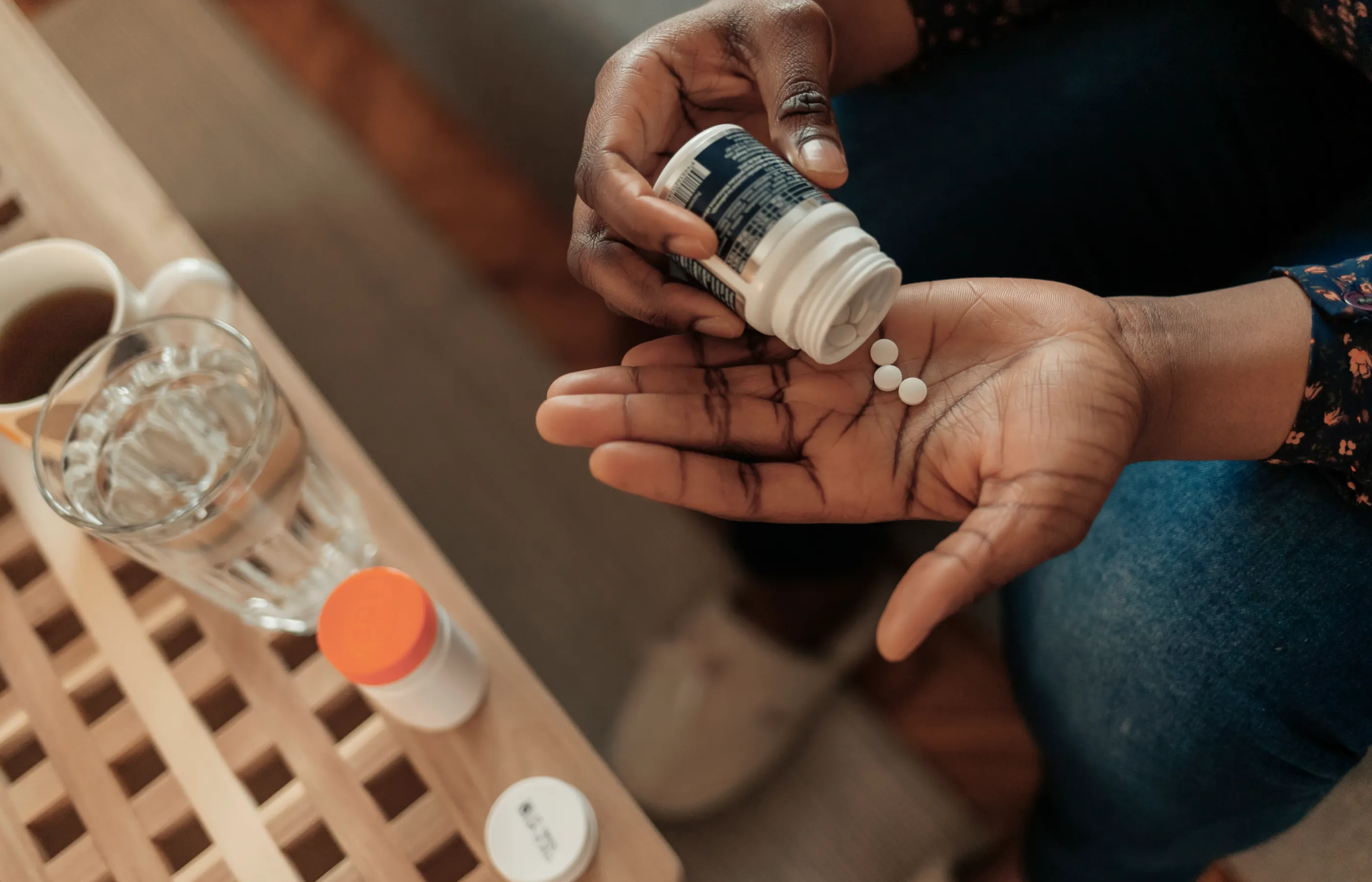 Patient is sitting on the couch with medication and glass of water placed on the coffee table. They are pouring medication in their hand. 