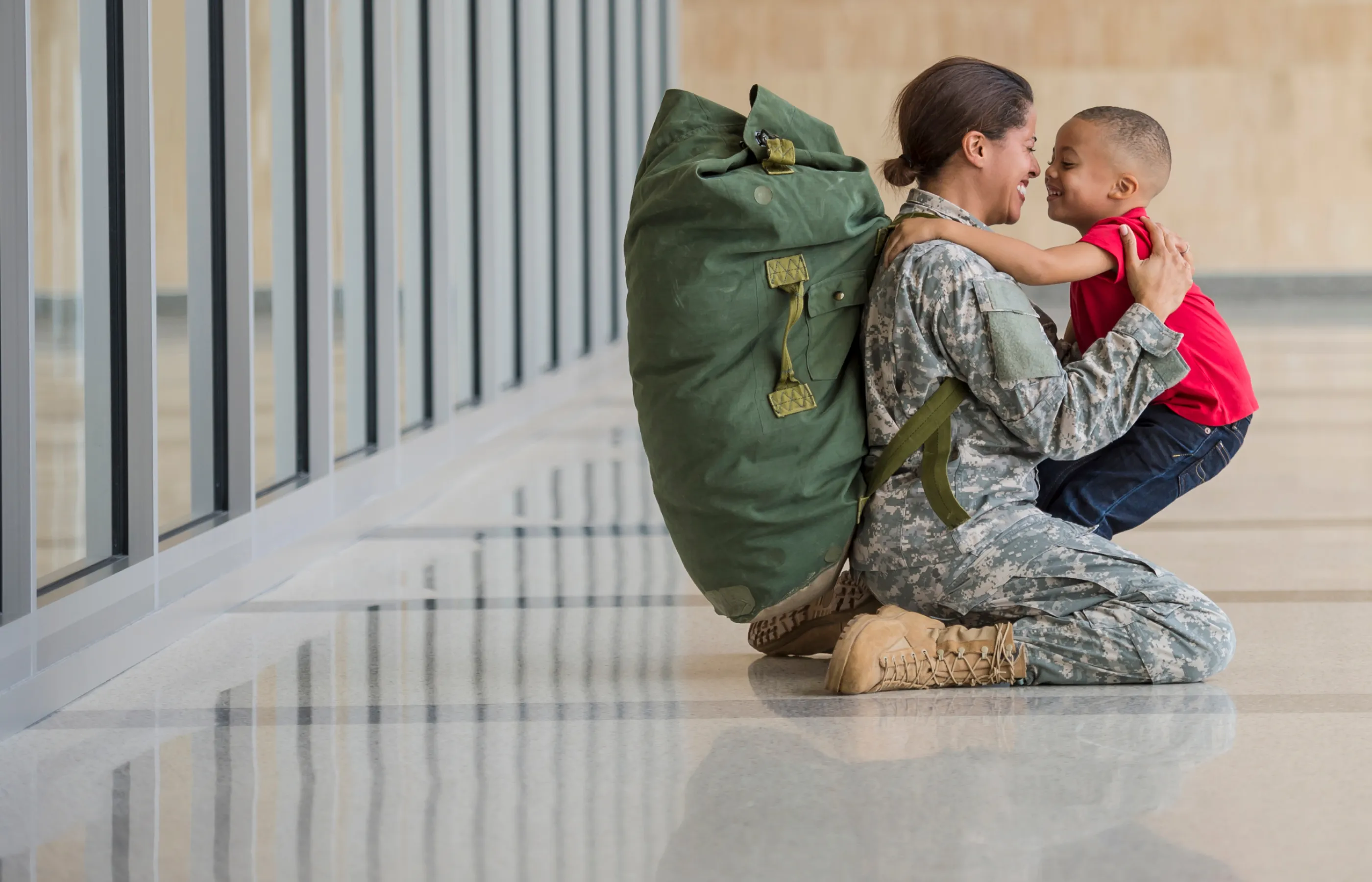 A woman is carrying a large military bag on her back while dressed in uniform. She is kneeling down on the floor as she hugs her young son. 