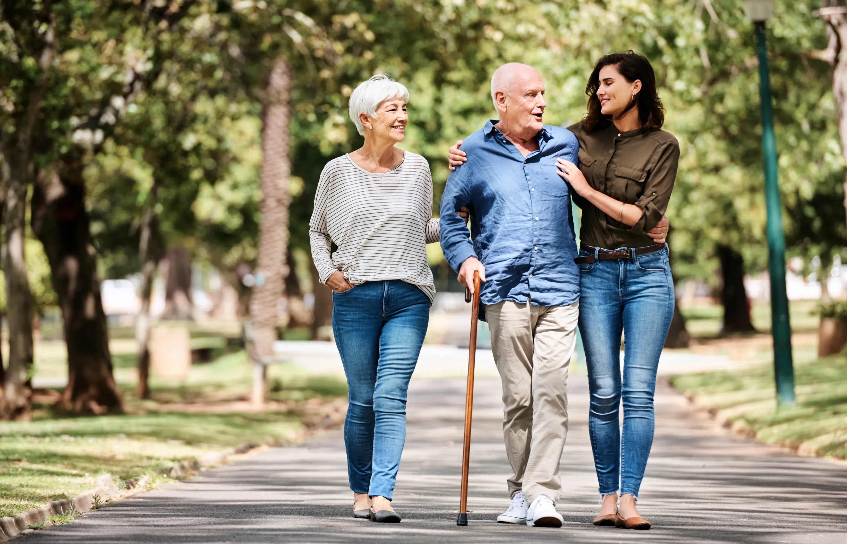 A senior man is walking at a park on a sunny day, with a walking cane in hand. He has his partner and adult daughter by his side. 