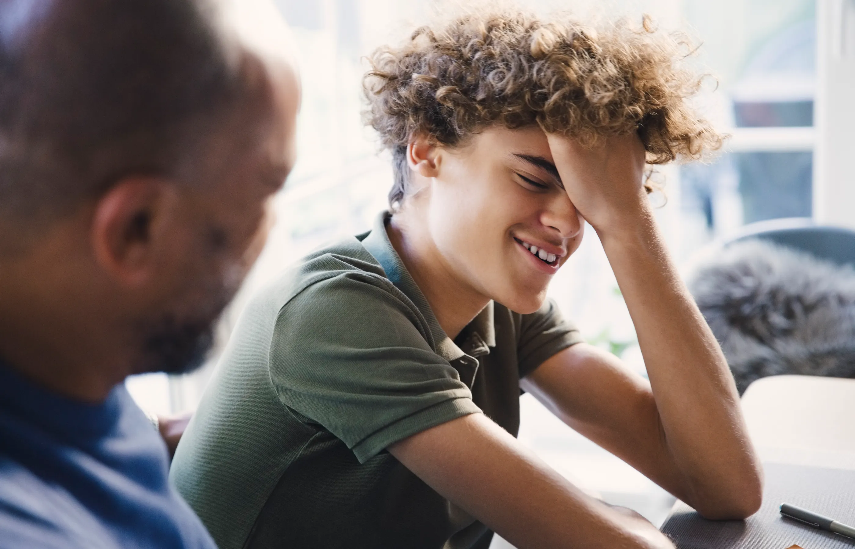 A teenage boy has his hand on his forehead as he smiles and talks with his dad. 