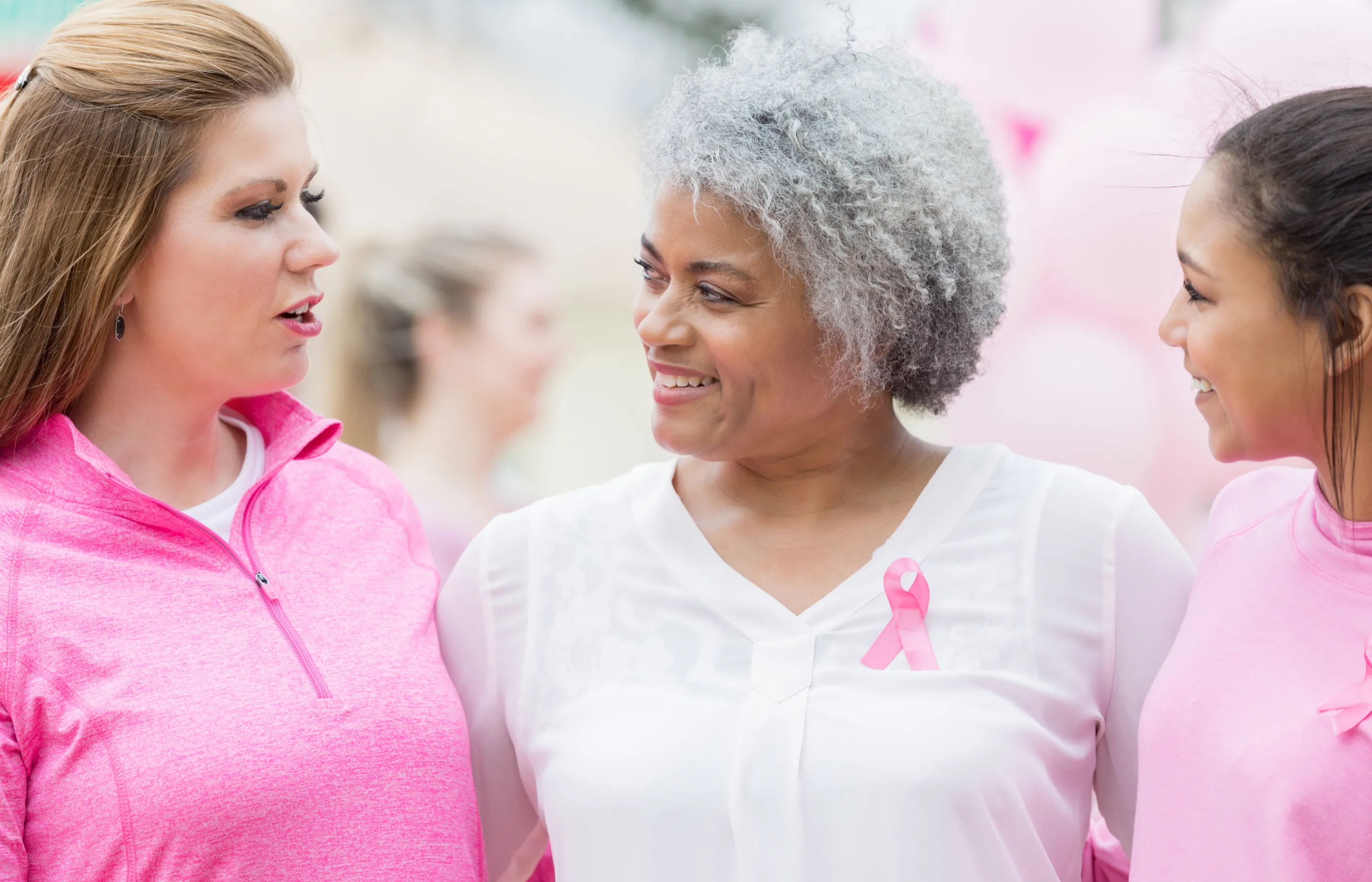 Three women are standing and together wearing white and pink. They are embracing each other as they prepare for a breast cancer event. 