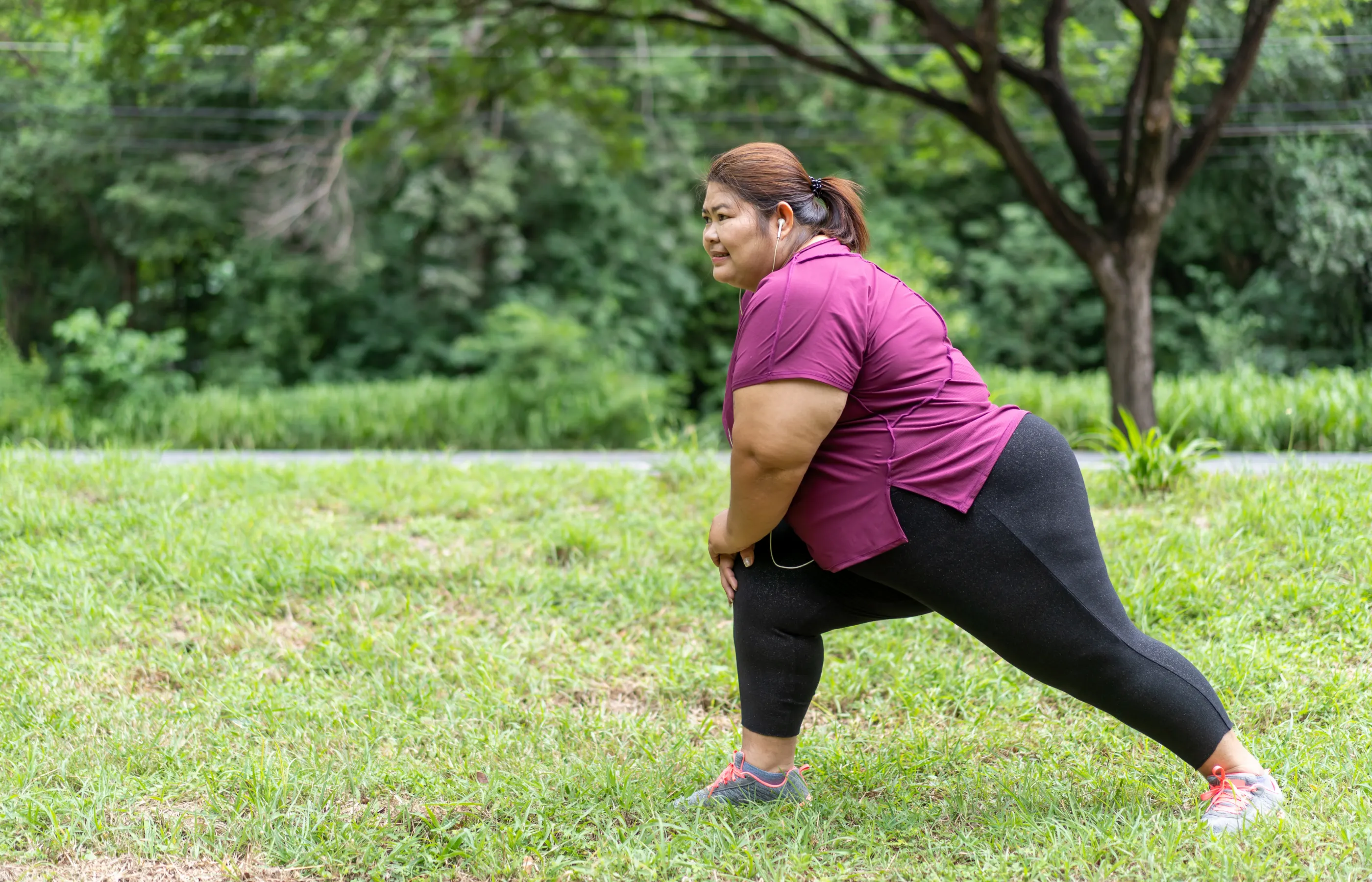 A woman is outdoors, stretching with headphones in her ear.
