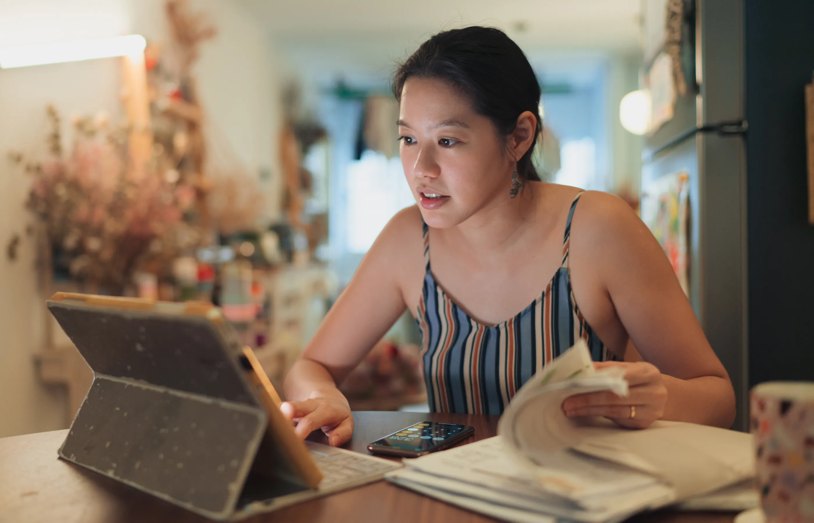 Woman sitting at her kitchen table with her tablet and a stack of bills. 