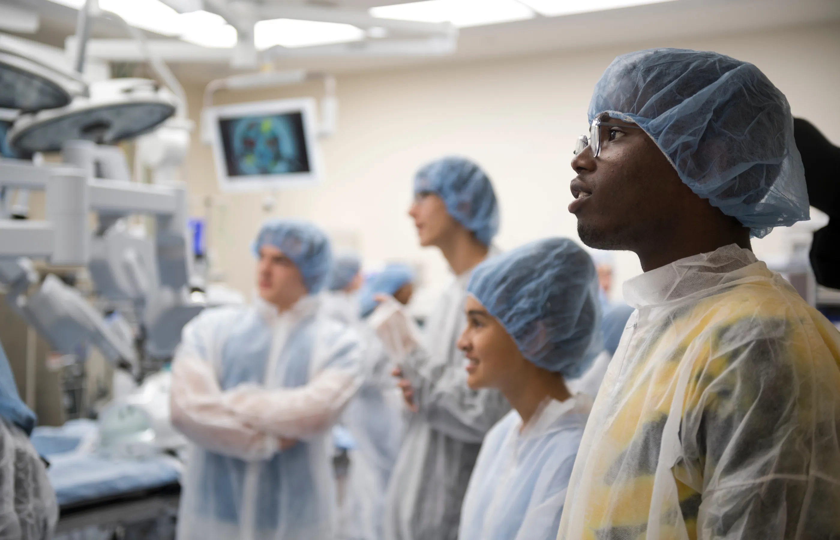 A group of students are wearing personal protective equipment (PPE) as they stand and observe in an operating room.   A group of students are wearing personal protective equipment (PPE) as they stand and observe in an operating room.