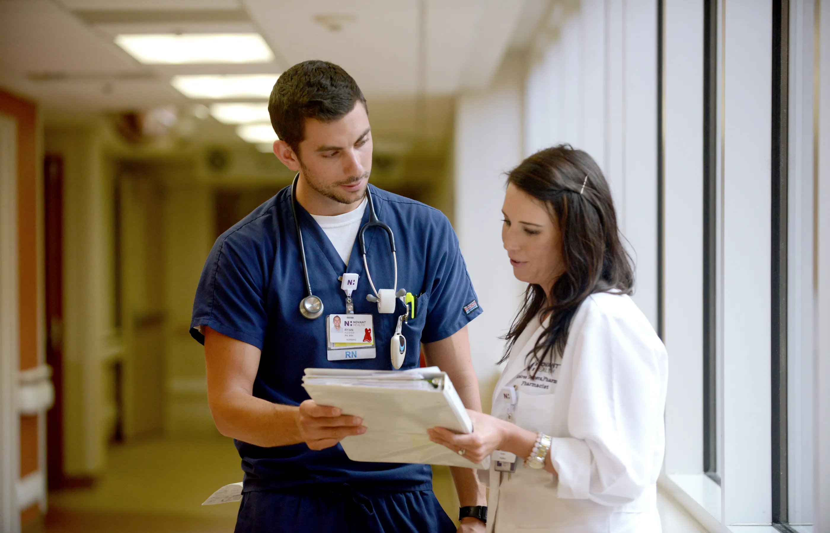 A Novant Health nurse and pharmacists are standing in a hall together, talking, and reviewing information. 