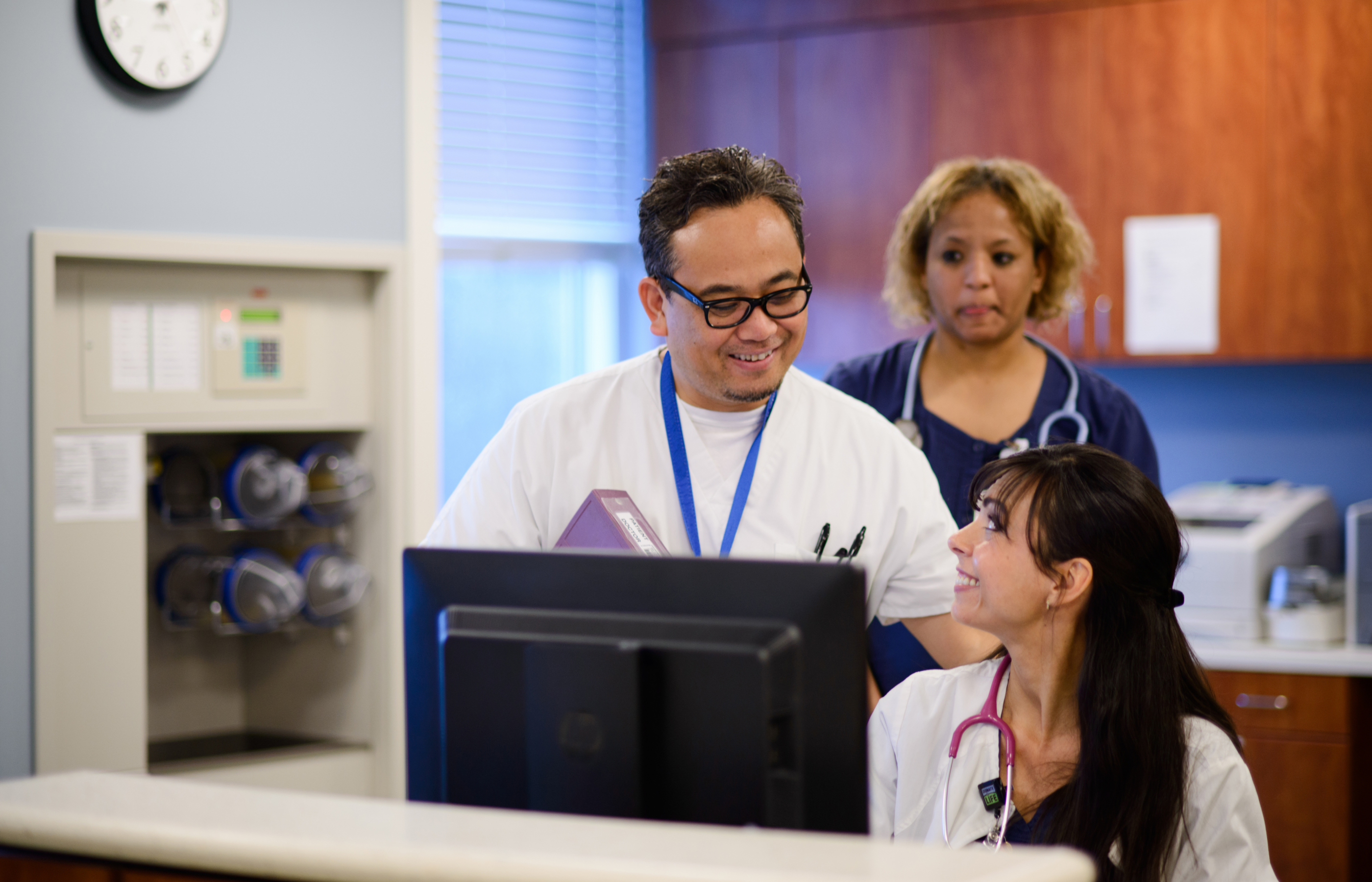 Three smiling nurses working at computer, talking in clinic office.