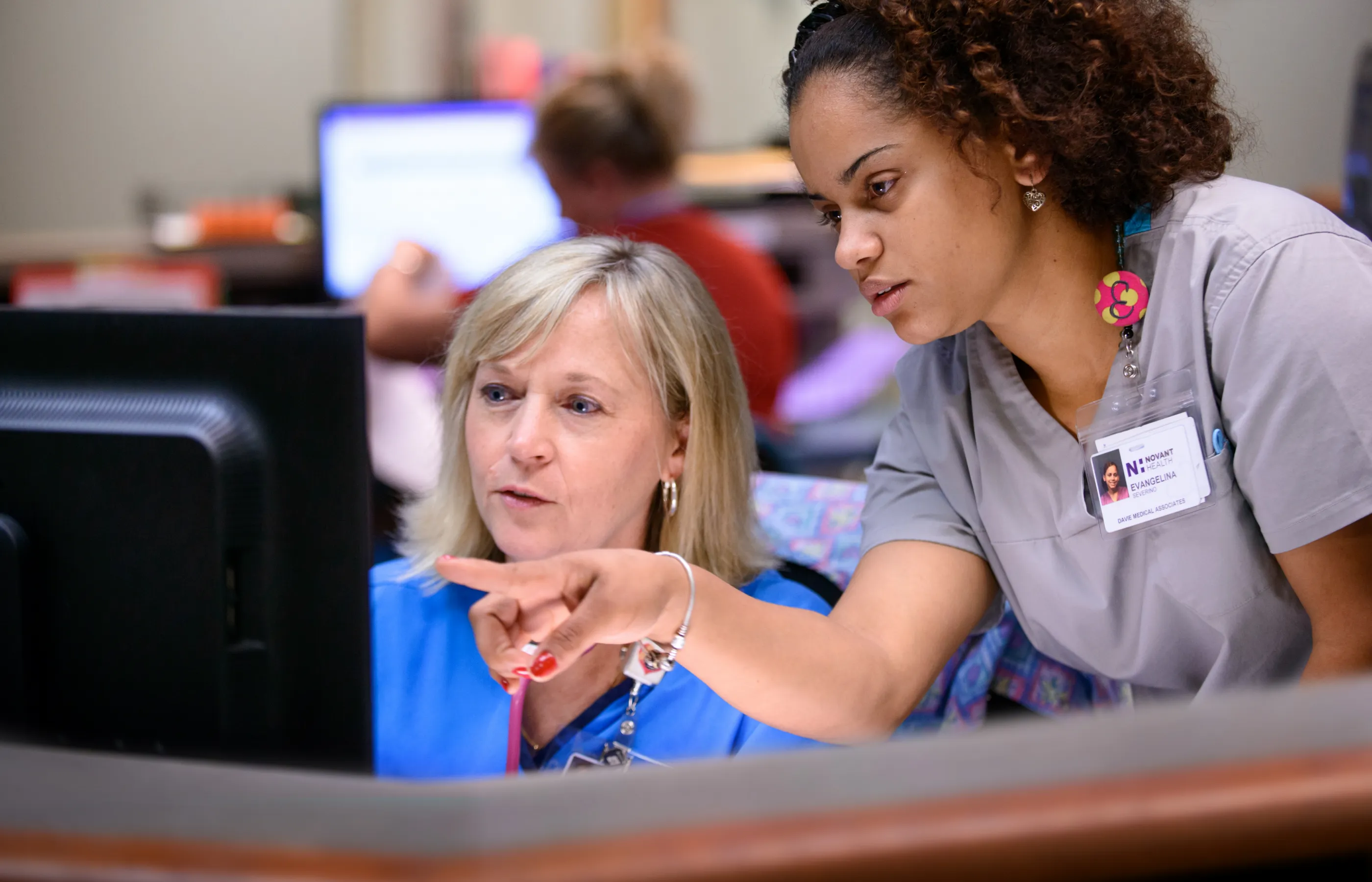 One Novant Health team member is sitting at a computer, the other is standing beside her and pointing at the computer screen. 