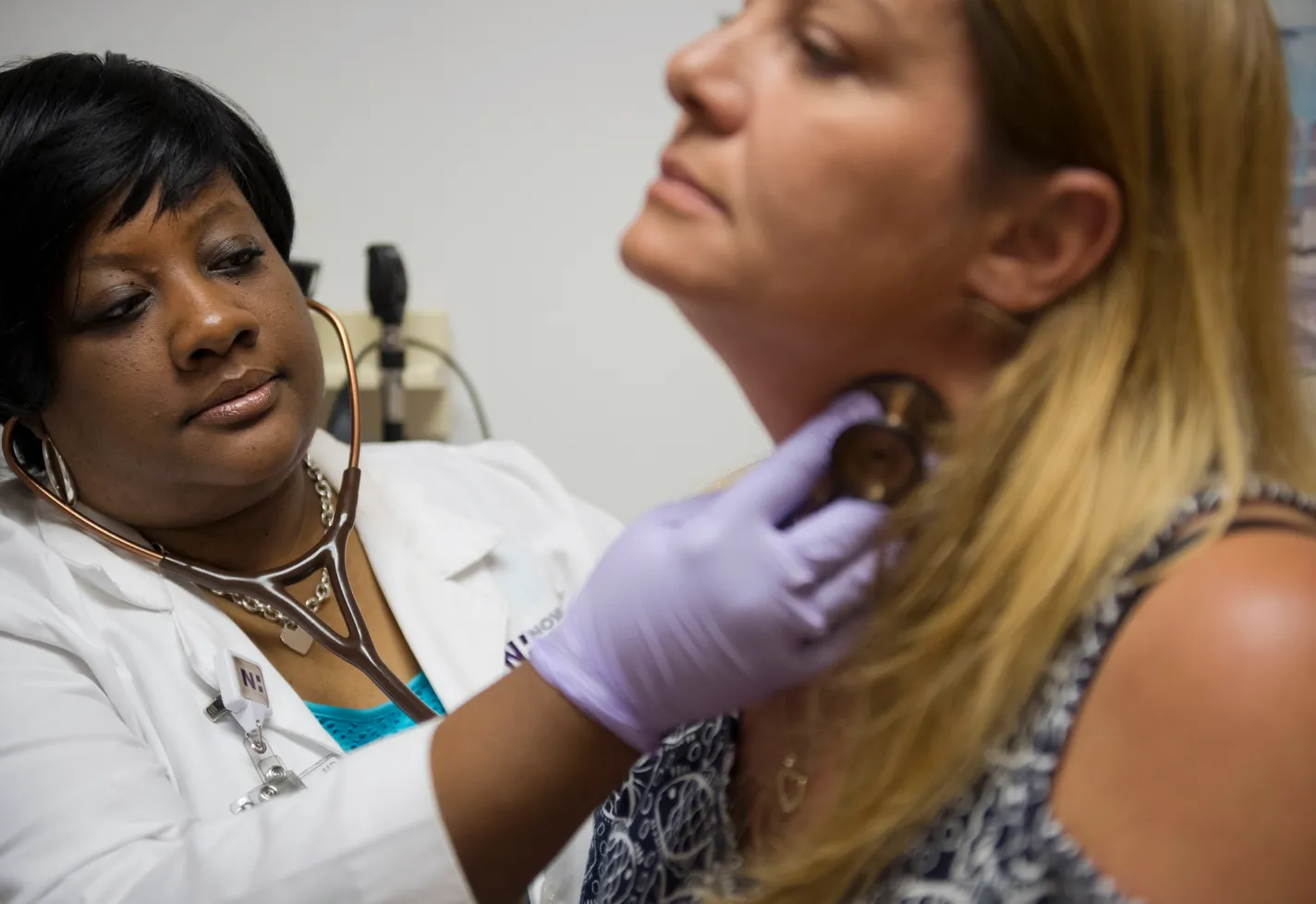 Dr. Thompson is in a clinic exam room with a patient. She is wearing a stethoscope and places it on the patients neck to listen. 