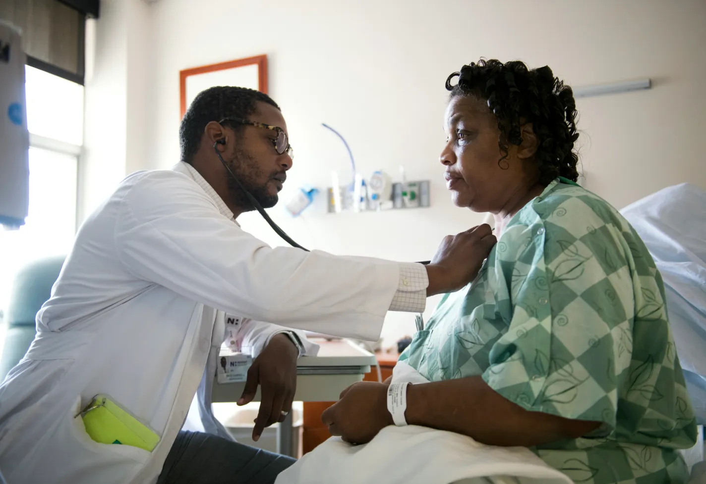 Dr. Campbell is sitting with a patient in their recovery room. With his stethoscope he listens to the patient's heart. 