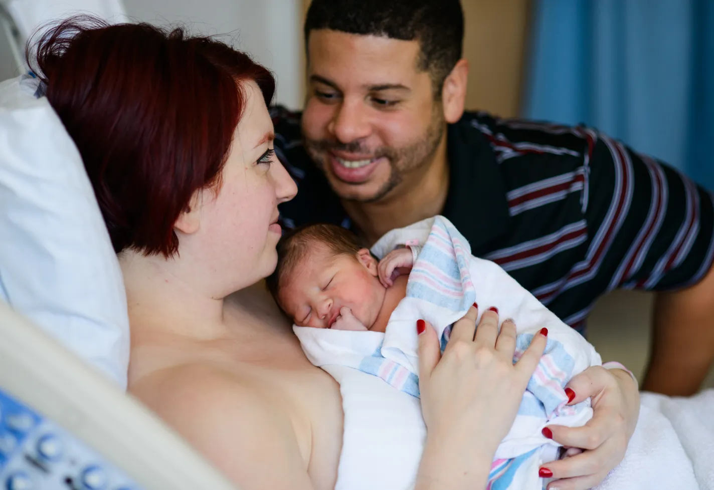 A woman is lying down in a hospital bed holding her newborn as her partner surrounds them both. 