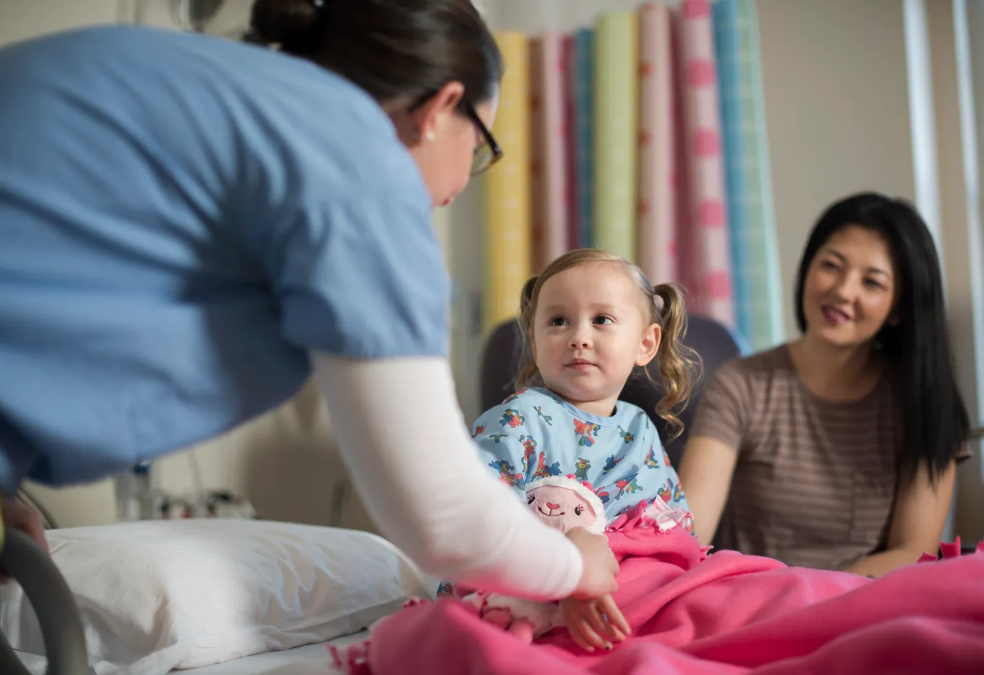 Young child sitting in a hospital bed with a stuffed animal. The childs mother is along side her as a Novant Health nurse approached them both. Young child sitting in a hospital bed with a stuffed animal. The childs mother is along side her as a Novant Health nurse approached them both.