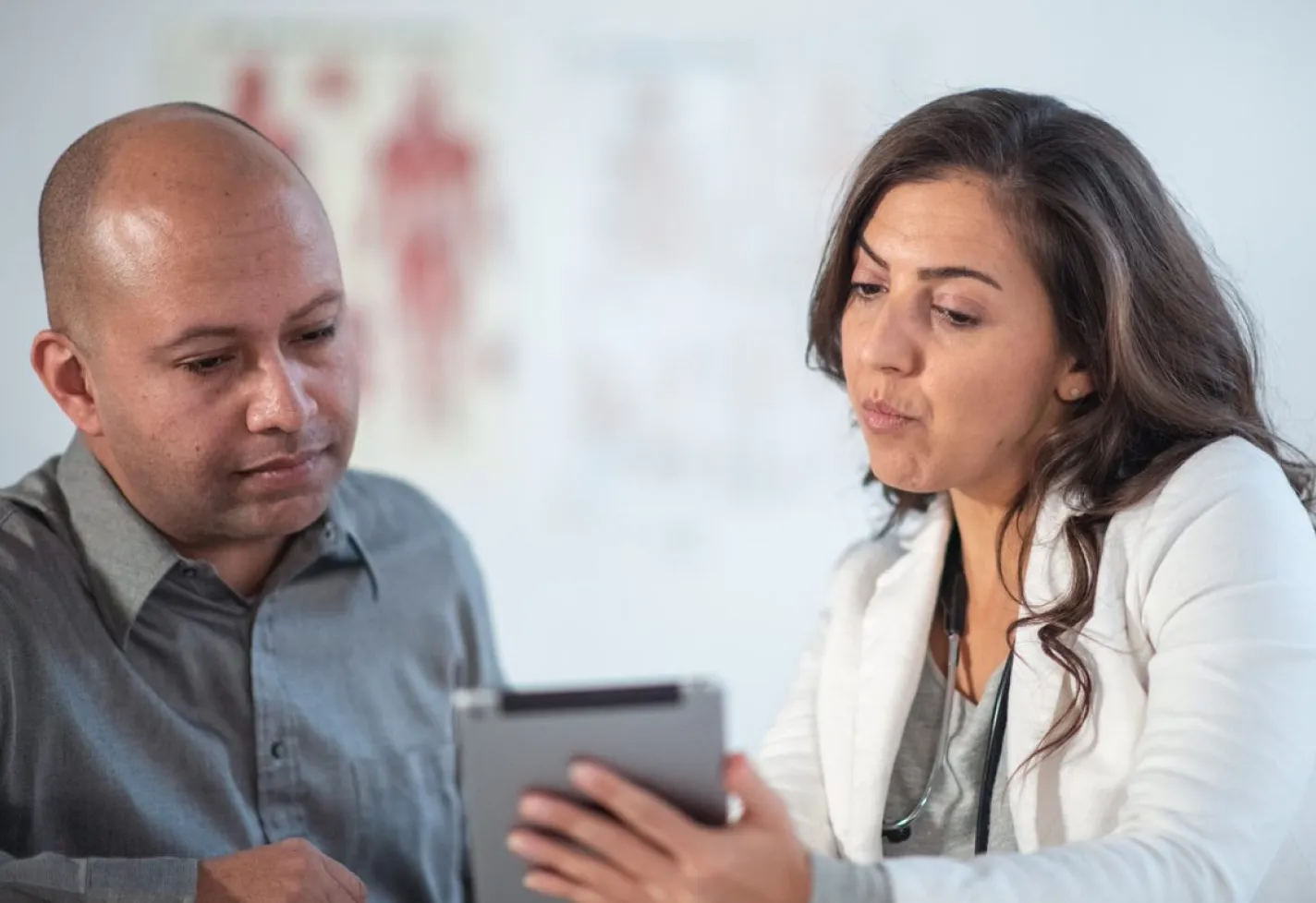 A male patient is sitting with a healthcare provider in lab coat. They are reviewing information on a smart tablet in an exam room.  