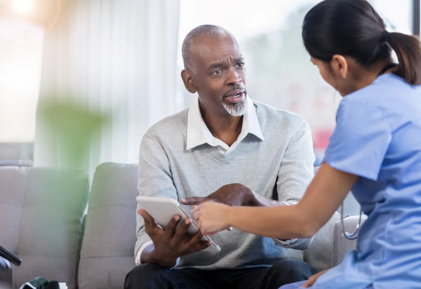 A male patient is sitting and talking with a nurse. Together, they are reviewing information on a tablet.