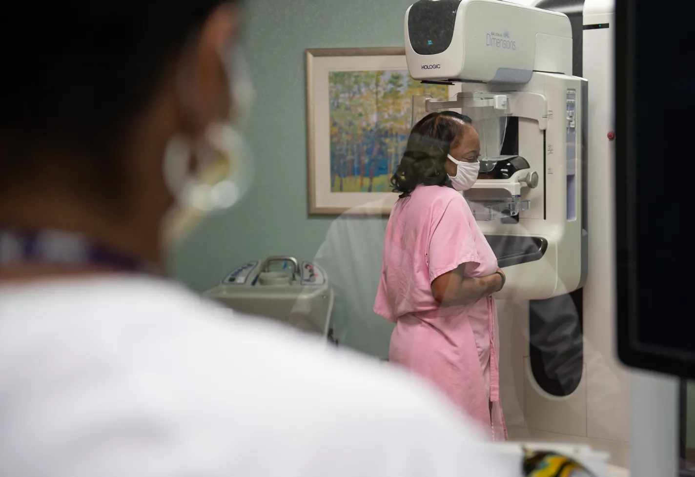 A Novant Health team member is standing behind a screen as a patient stands in a pink gown to have her mammogram screening. A Novant Health team member is standing behind a screen as a patient stands in a pink gown to have her mammogram screening.
