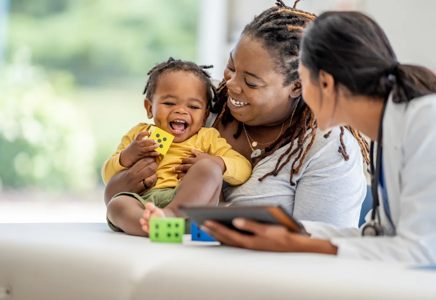 A mom, her toddler, and a provider holding a tablet smile as the baby plays with dice blocks A mom, her toddler, and a provider holding a tablet smile as the baby plays with dice blocks