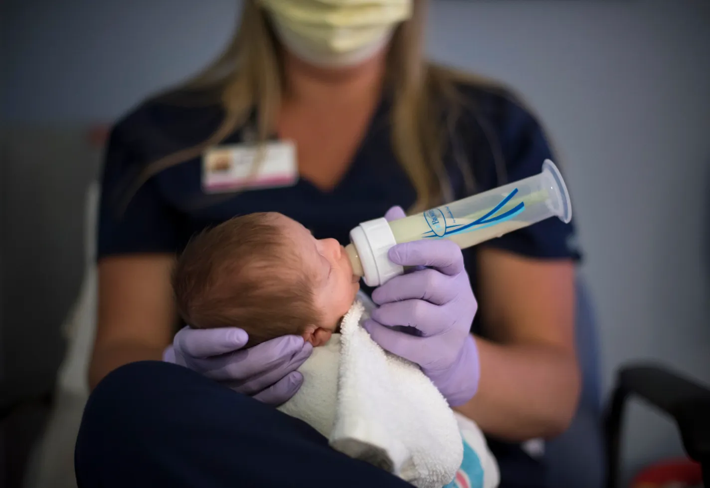 A Novant Health nurse is wearing gloves as she holds a newborn baby and feeds them a bottle. 