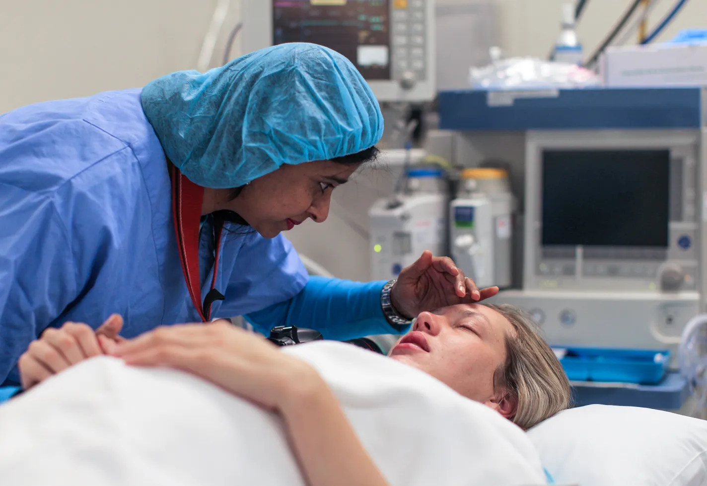 A pregnant patient is lying down in an operating room as a healthcare provider leans in close to talk to her. As the healthcare provider is talking, she hold one of the patients hands. With the other she gently brushes the hair out of her face. 