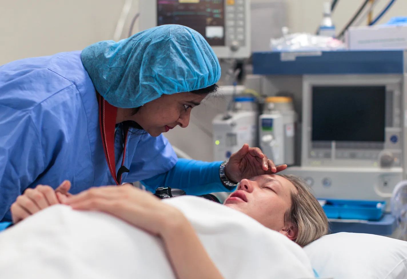 A pregnant patient is lying down in an operating room as a healthcare provider leans in close to talk to her. As the healthcare provider is talking, she hold one of the patients hands. With the other she gently brushes the hair out of her face. A pregnant patient is lying down in an operating room as a healthcare provider leans in close to talk to her. As the healthcare provider is talking, she hold one of the patients hands. With the other she gently brushes the hair out of her face.