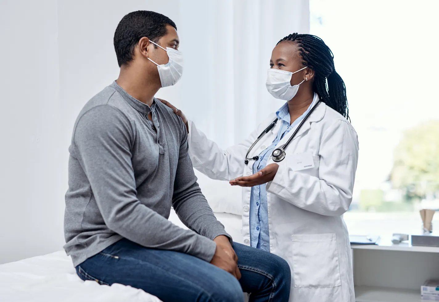 A doctor and patient are both wearing masks during their visit. The patient is sitting on the exam table as the provider is talking and resting her hand on his shoulder. 