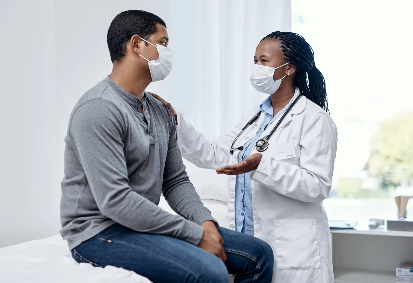 A doctor and patient are both wearing masks during their visit. The patient is sitting on the exam table as the provider is talking and resting her hand on his shoulder. A doctor and patient are both wearing masks during their visit. The patient is sitting on the exam table as the provider is talking and resting her hand on his shoulder.