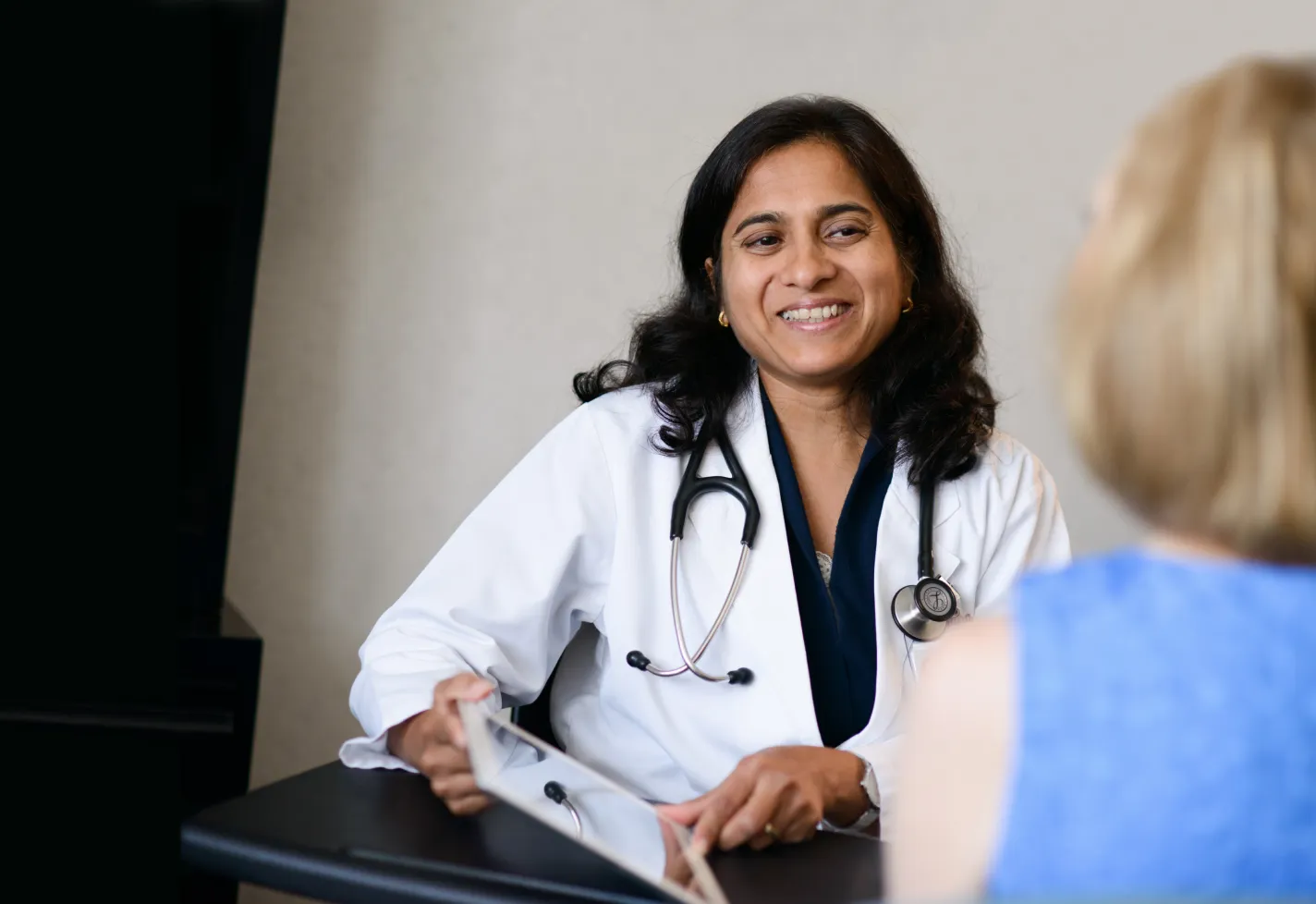 A health care provider is sitting with a patient talking as she wears a lab coat and stethoscope. 