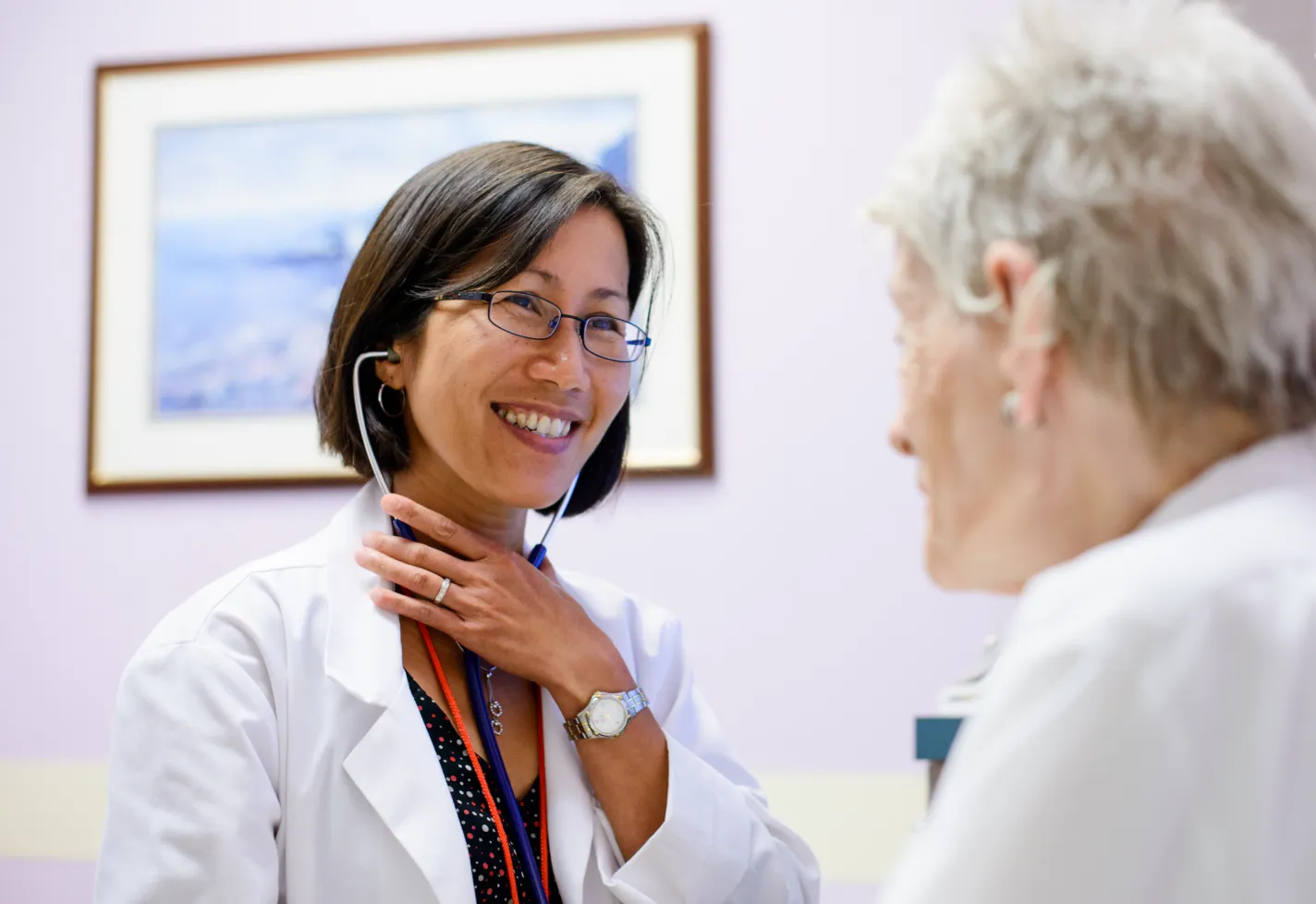 A doctor is sitting with a senior patient in an exam room. The doctor is wearing a stethoscope as she smiles at the patient. 