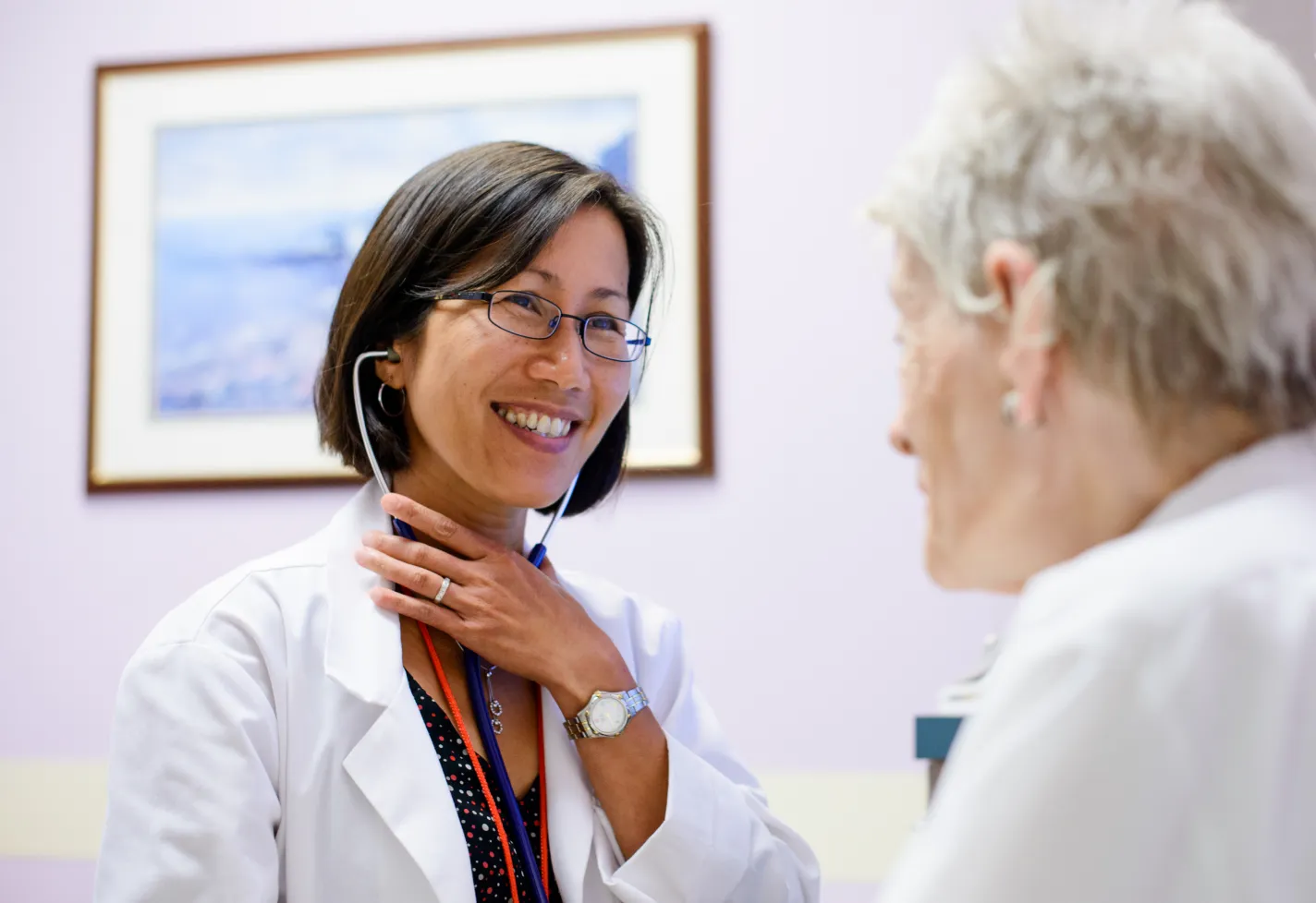 A doctor is sitting with a senior patient in an exam room. The doctor is wearing a stethoscope as she smiles at the patient. A doctor is sitting with a senior patient in an exam room. The doctor is wearing a stethoscope as she smiles at the patient.