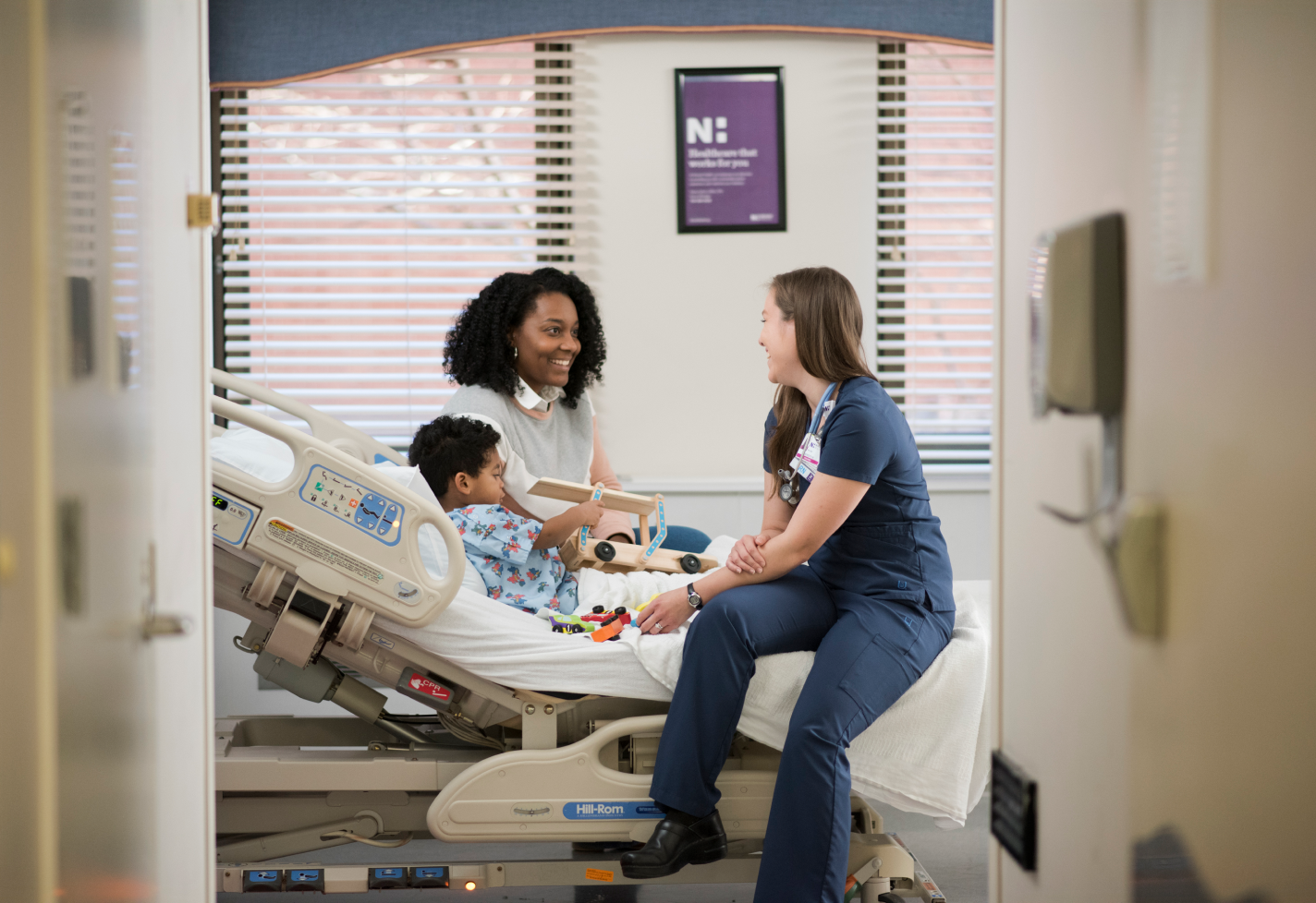 A Novant Health nurse is sitting on a hospital bed as she talks with a young patient and their mom. 