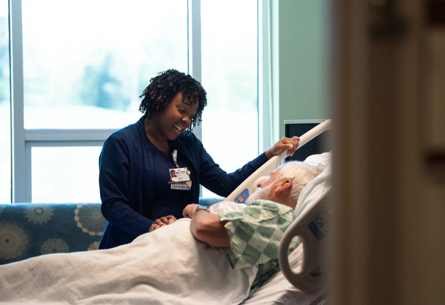 A Novant Health nurse is standing at the bedside of a patient talking with the patient. 