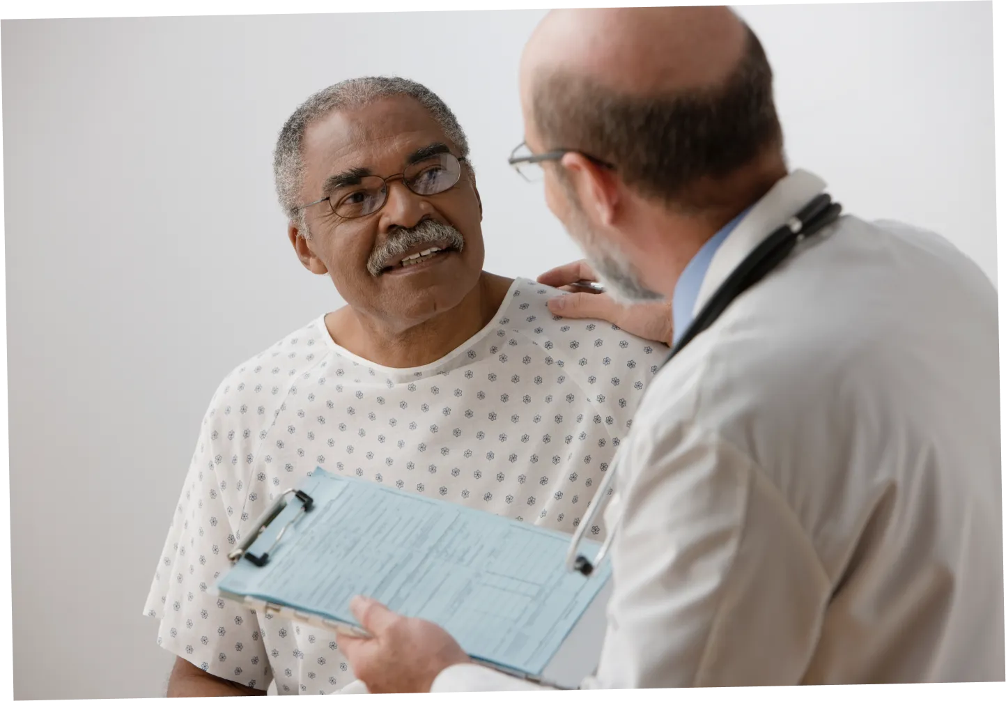 A senior man is talking with his doctor as he sits on the exam table in a gown. The doctor has placed his hand on the patients shoulder.